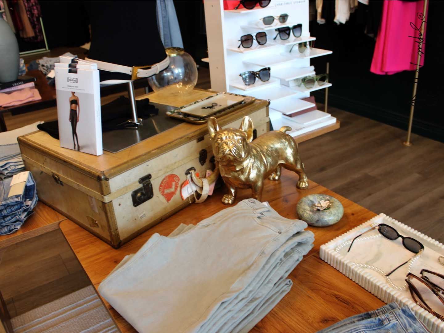 Retail display with vintage suitcase, gold dog figurine, folded clothes, and sunglasses on a wooden table