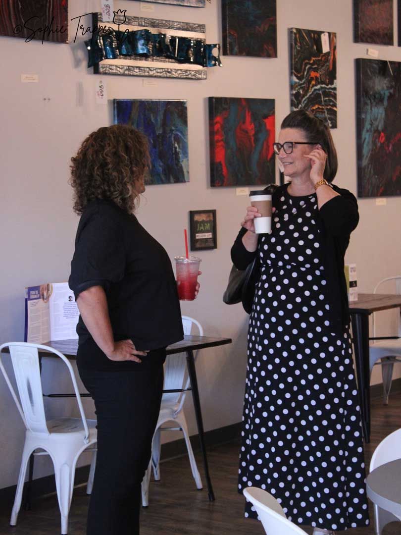 Two people talking in an art gallery, one holding a drink and smiling in a polka-dot dress.