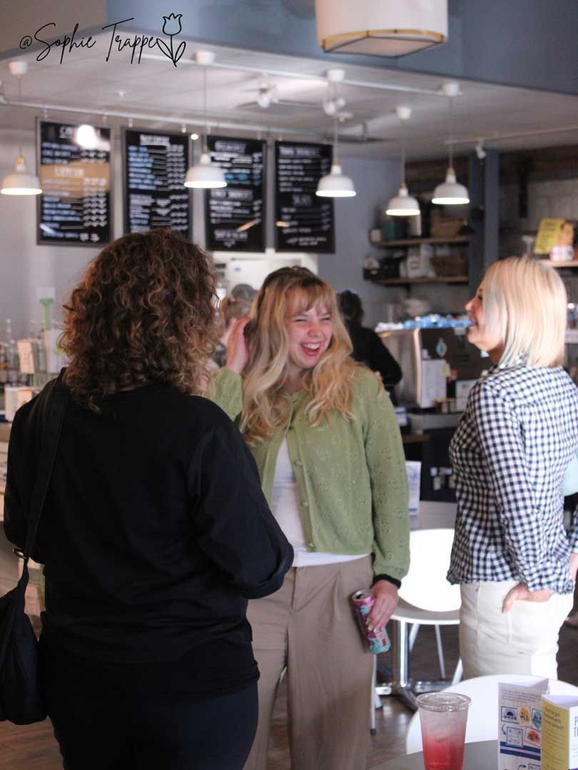 Three people chatting inside a bright café with menu boards and hanging pendant lights.