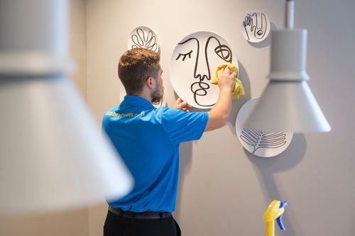 Man in blue shirt cleaning a wall-mounted art piece with a yellow cloth. Gray wall, white lamps.
