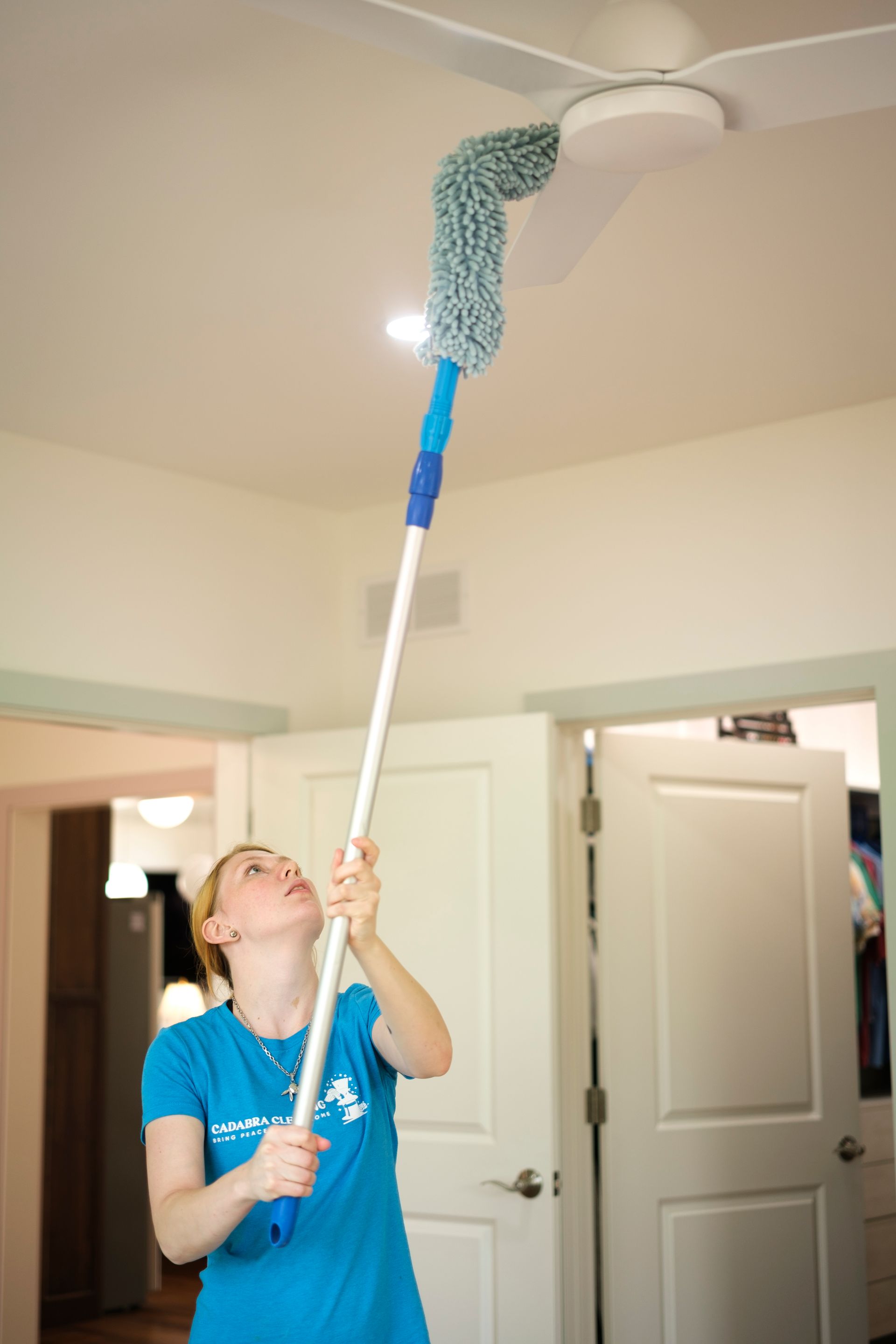 Woman in blue shirt cleaning a ceiling fan with an extendable duster indoors.
