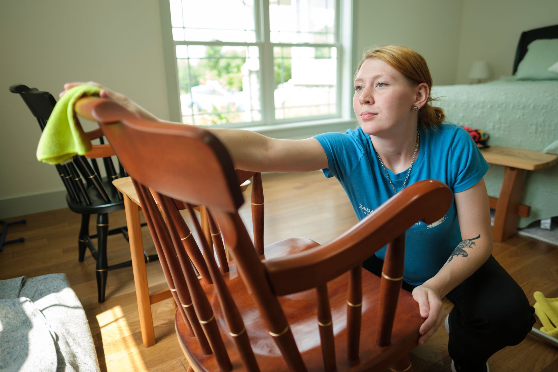 Woman cleaning wooden chair with a yellow cloth in a sunlit room.