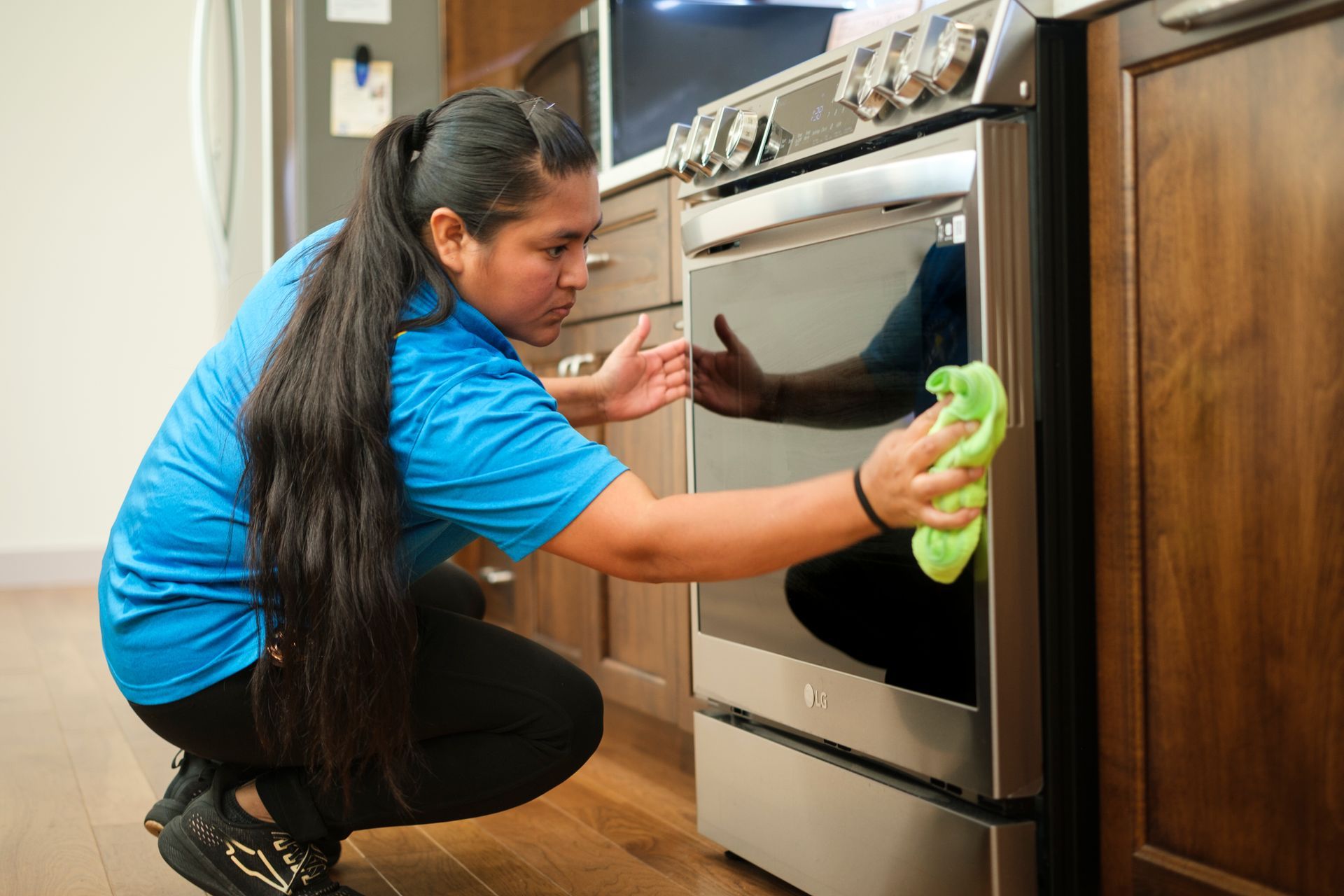 Woman in blue shirt cleaning a stainless steel oven door with a green cloth in a kitchen.