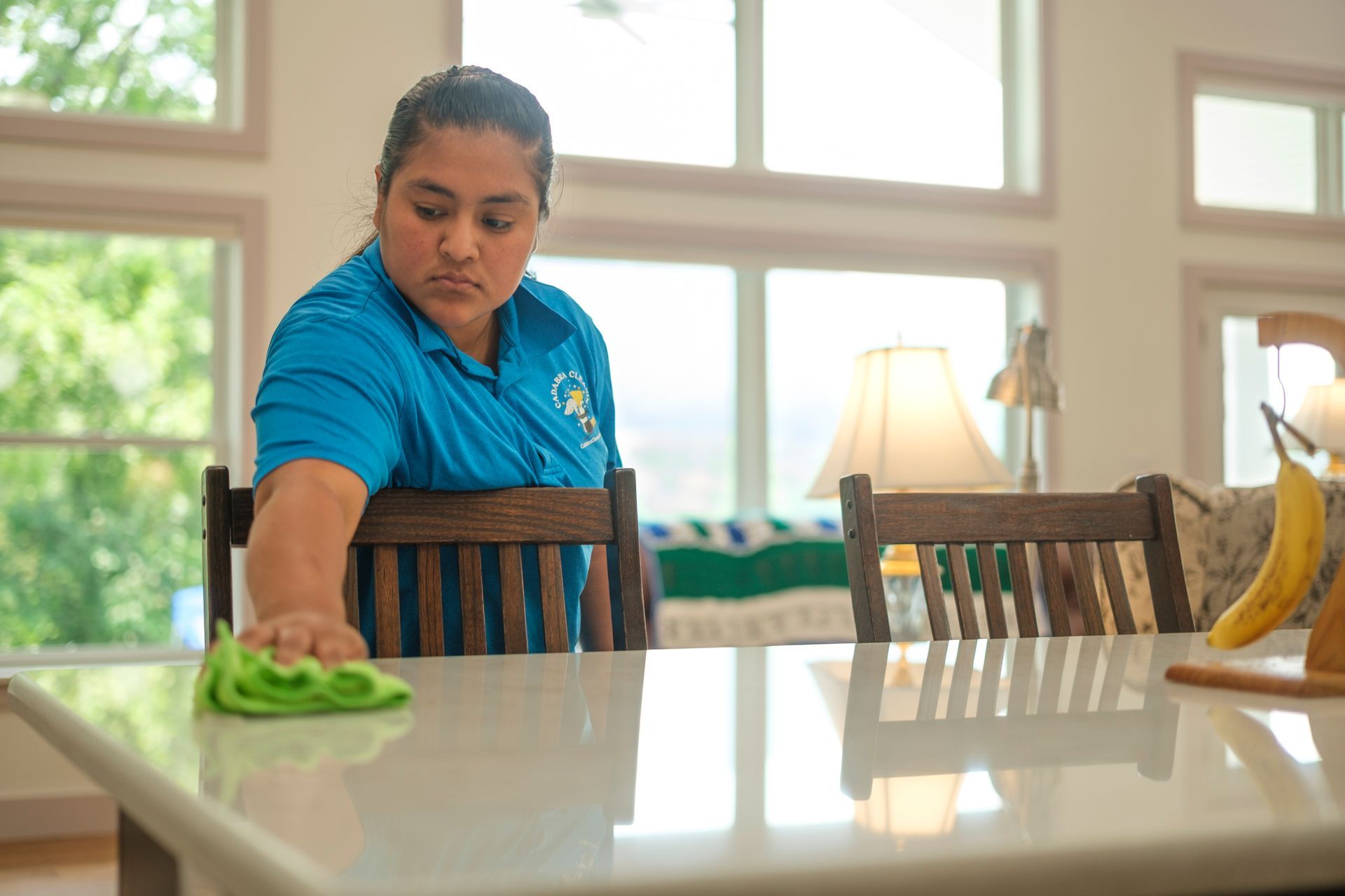 Woman in blue shirt cleaning a white counter in a bright room with large windows.