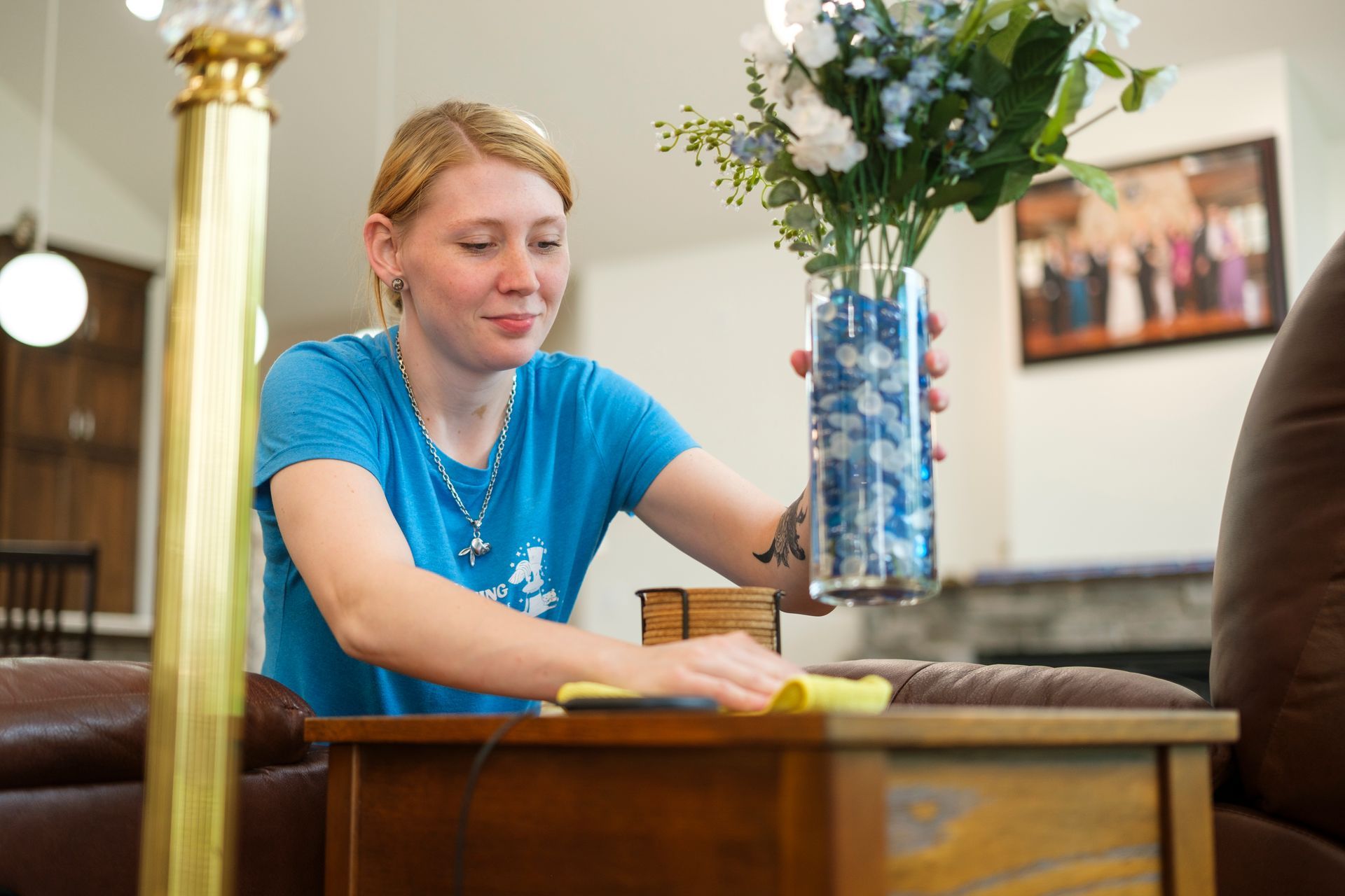 Woman cleaning a wooden table, holding a vase of flowers, in a living room.