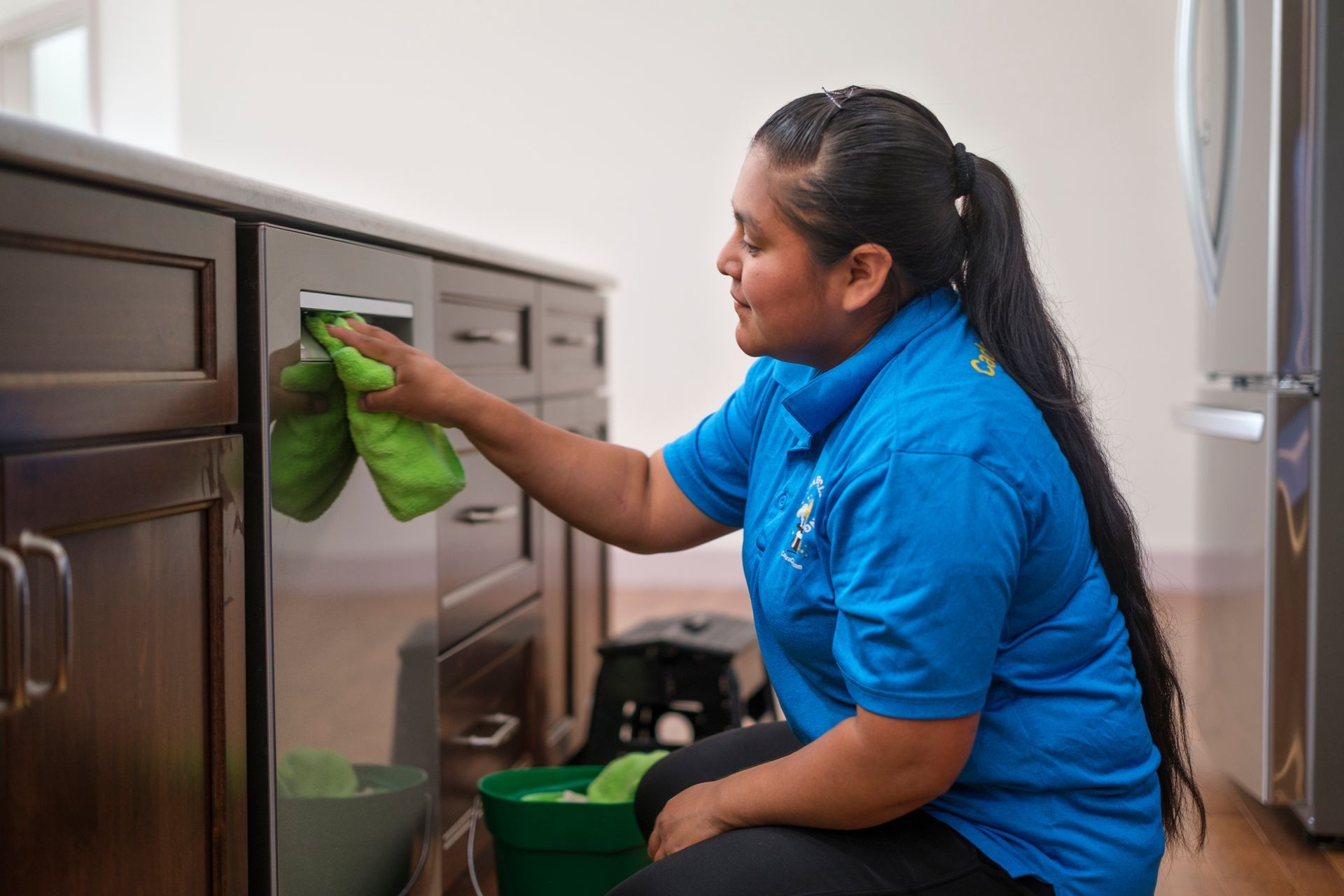 Woman cleaning a dark wooden cabinet in a kitchen with a green cloth.