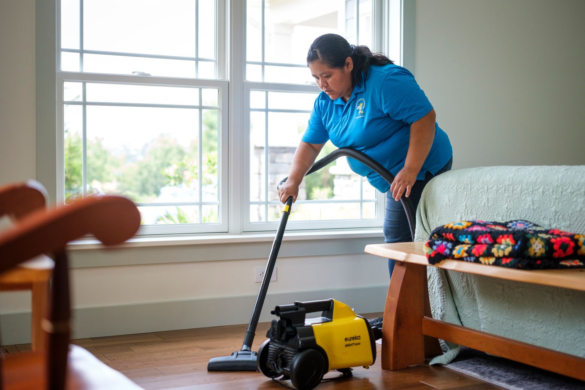 Woman in blue shirt vacuums a wood floor in front of a window. Vacuum cleaner is on the floor.