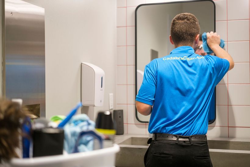 Man in blue shirt cleaning a bathroom mirror. He holds a blue cloth. White tiles and sink are in the background.