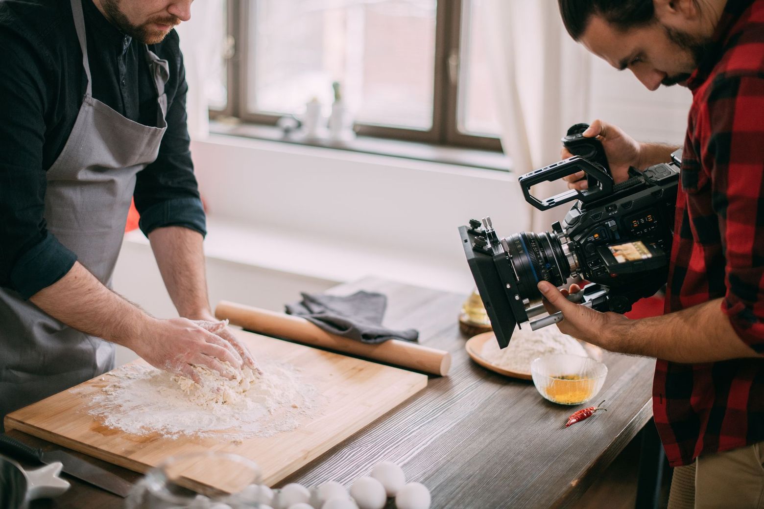 A man is rolling dough on a wooden cutting board while another man takes a picture with a camera.