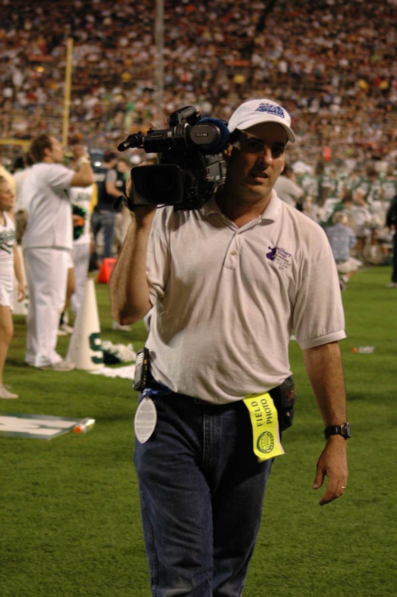 A man carrying a camera on a field with a tag that says ' o ' on it