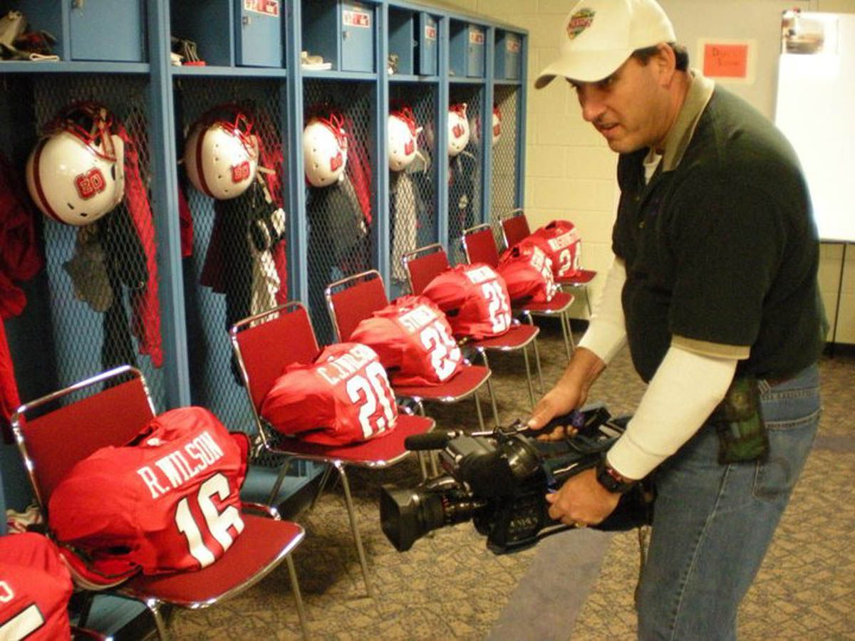 A man is holding a camera in a locker room with a wilson jersey on a chair
