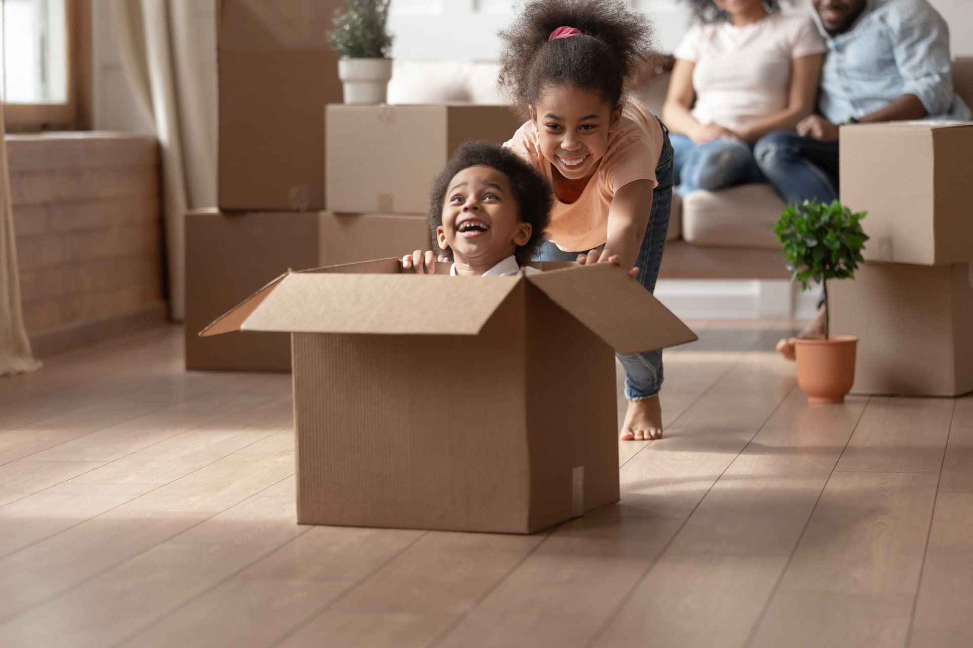 A boy and a girl are playing in a cardboard box in a living room.