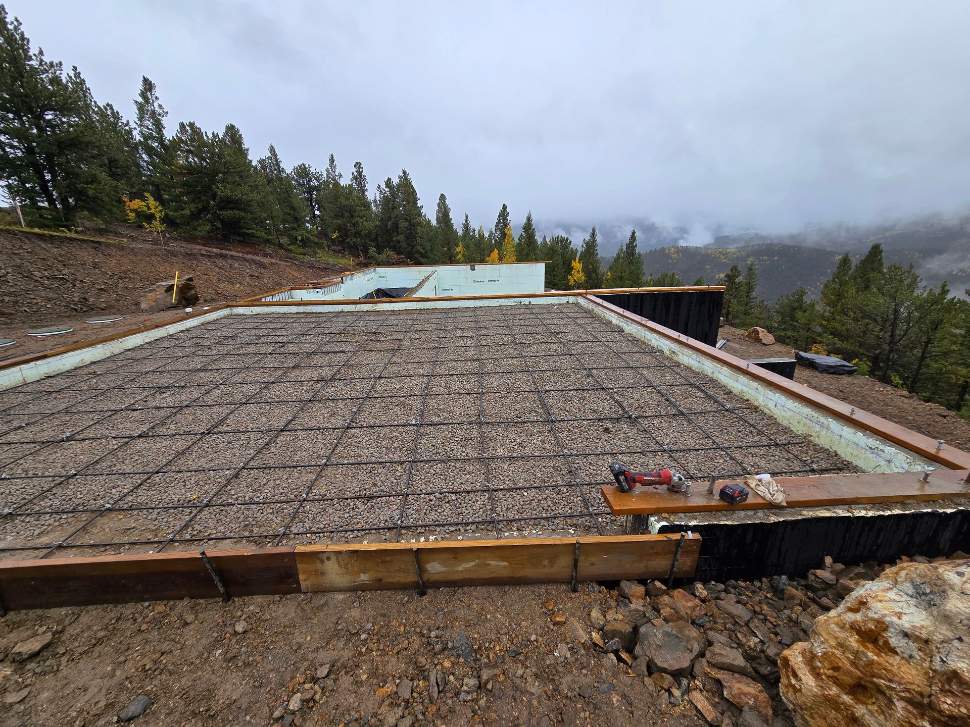 Large rebar foundation slab under construction on a hillside, with cloudy sky and pine trees nearby