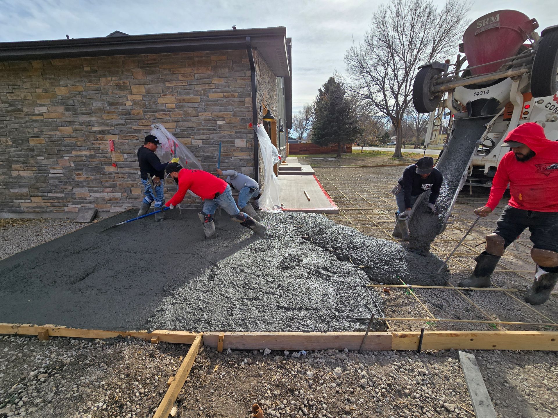 Workers pouring concrete beside a brick building, with a cement mixer and forms in place