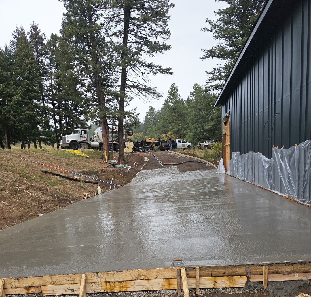 Fresh concrete driveway beside a dark metal building on a rainy construction site with trees and equipment nearby