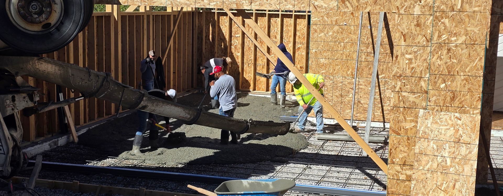 Construction workers pouring concrete near a wooden foundation wall, with a truck and gravel in the foreground