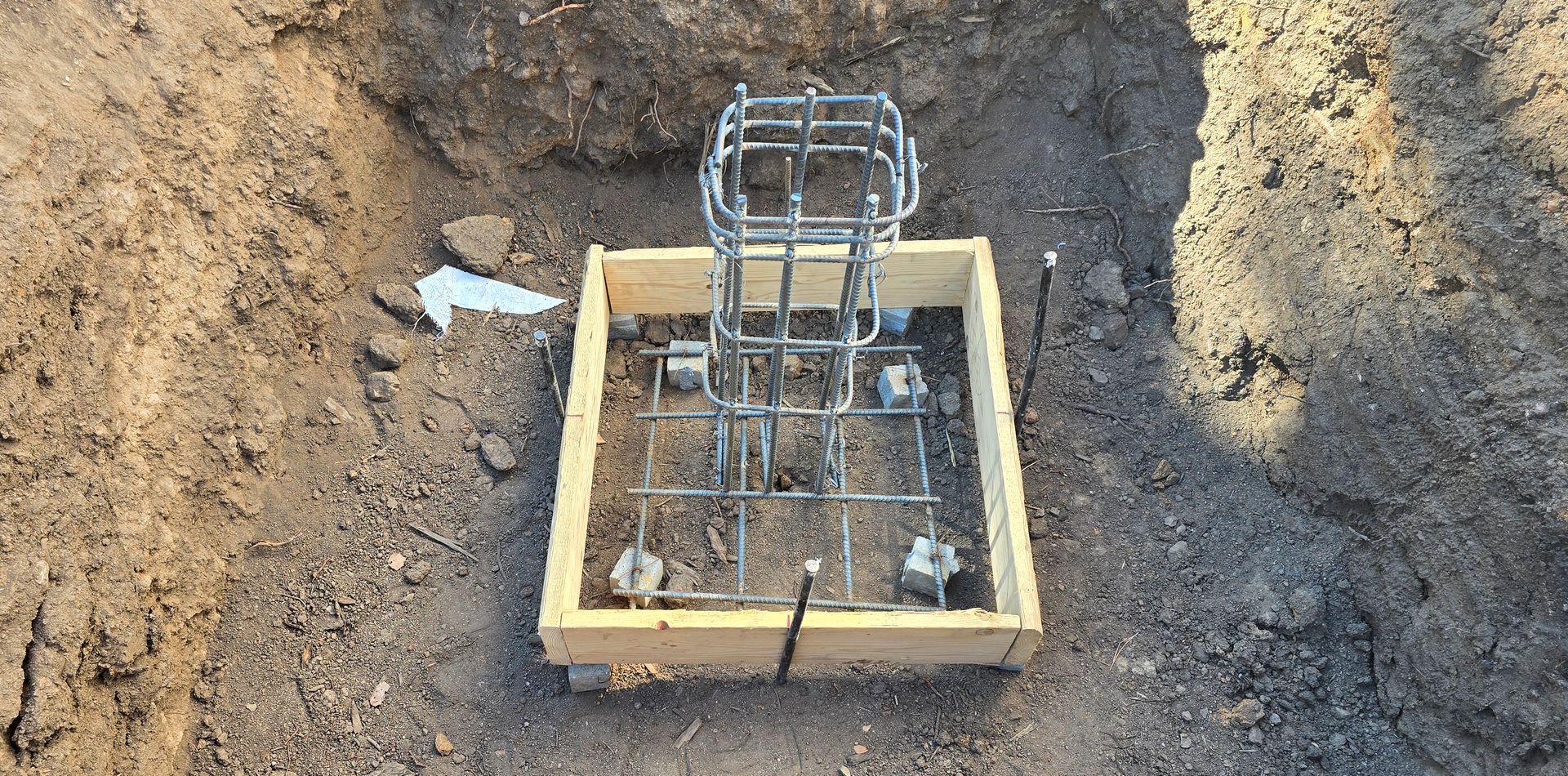 Wooden formwork around a rebar column footing in a dirt excavation pit.