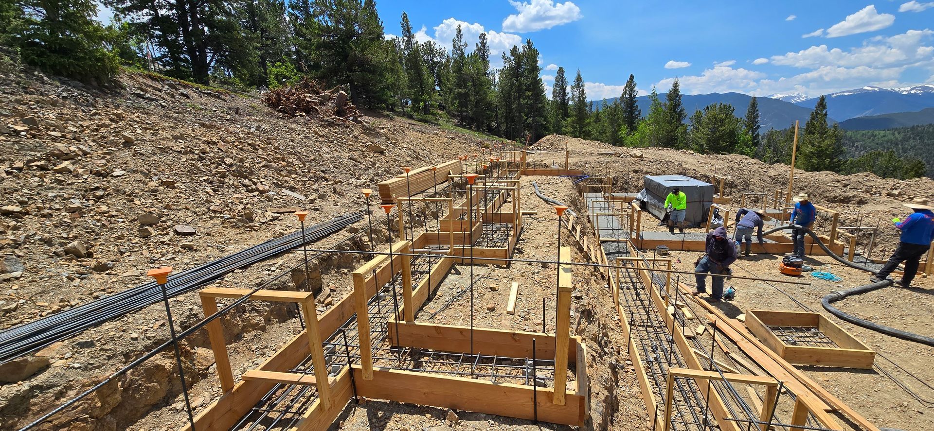Construction site with wooden foundation frames, workers, and blue sky on a rocky mountainside