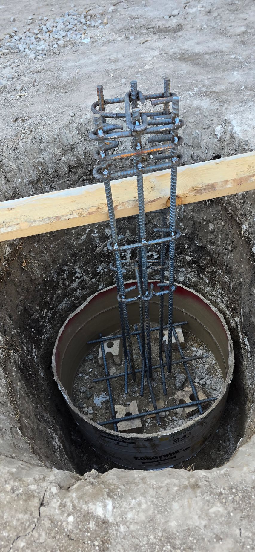 Steel rebar cage in a circular foundation hole at a construction site, with a wooden beam across it.