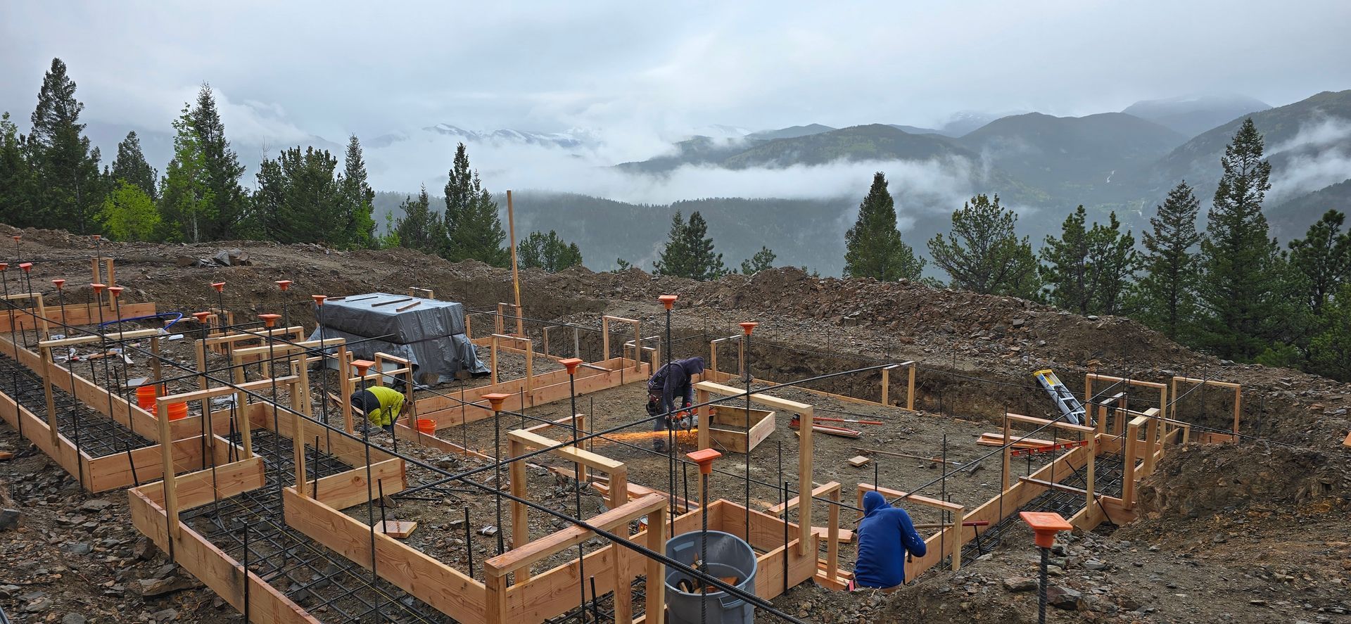 Construction site with workers framing a foundation in a muddy field, with mountains and cloudy sky in the background