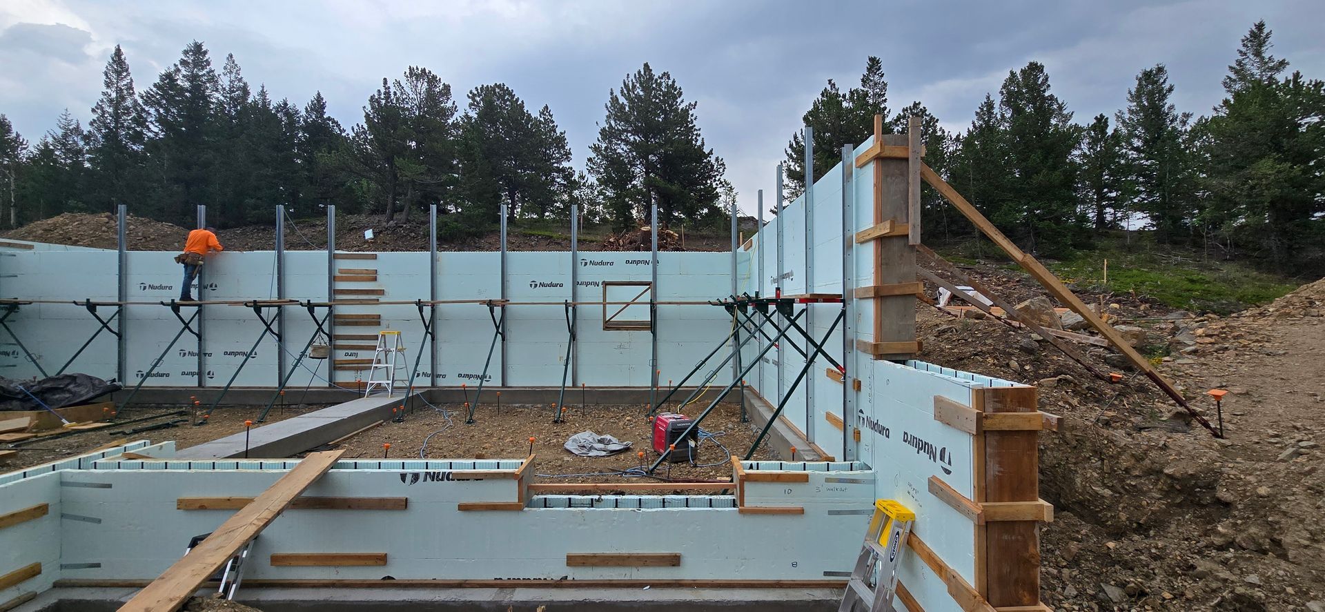 Construction site with concrete foundation walls, scaffolding, and lumber framing in a wooded area