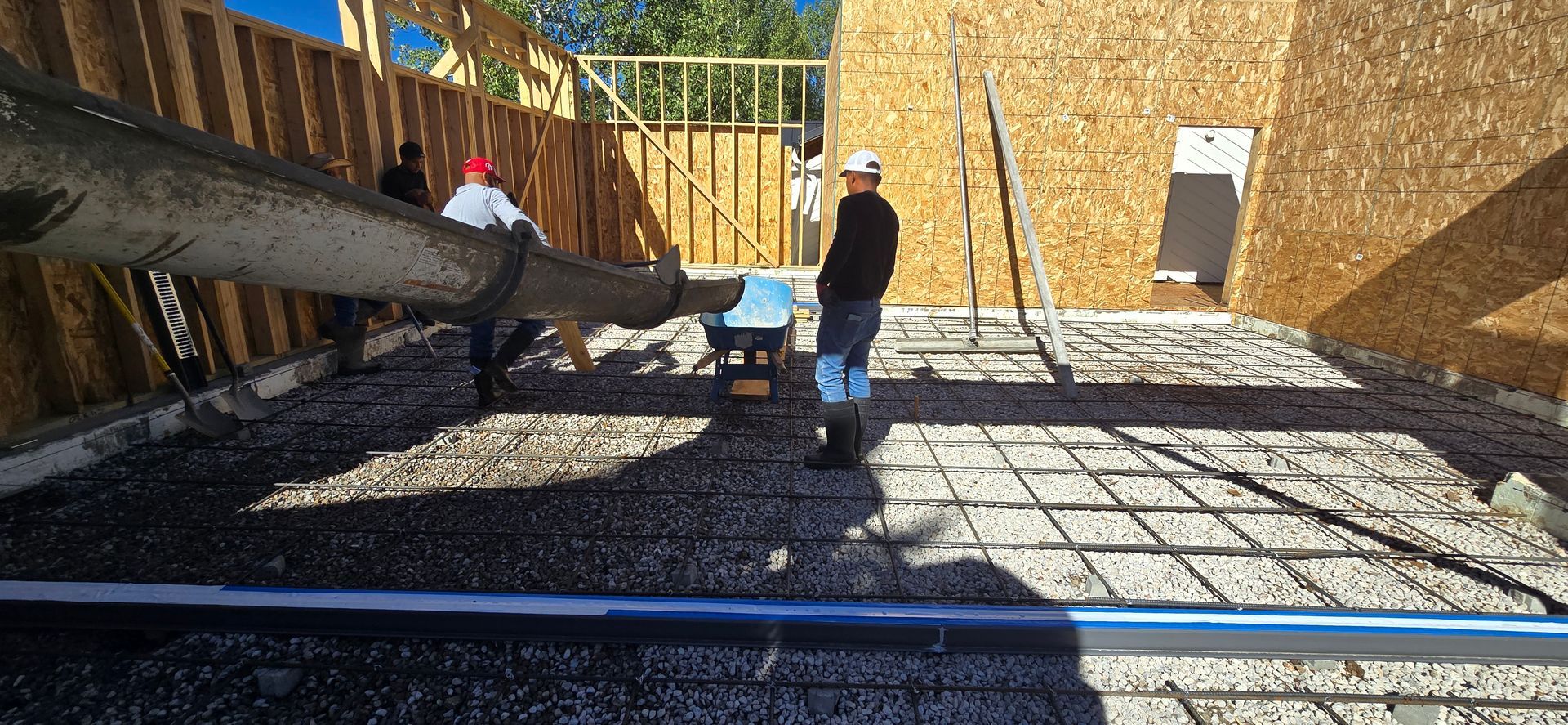 Two workers on a rooftop, one spraying sealant beside a plywood wall in bright sunlight.