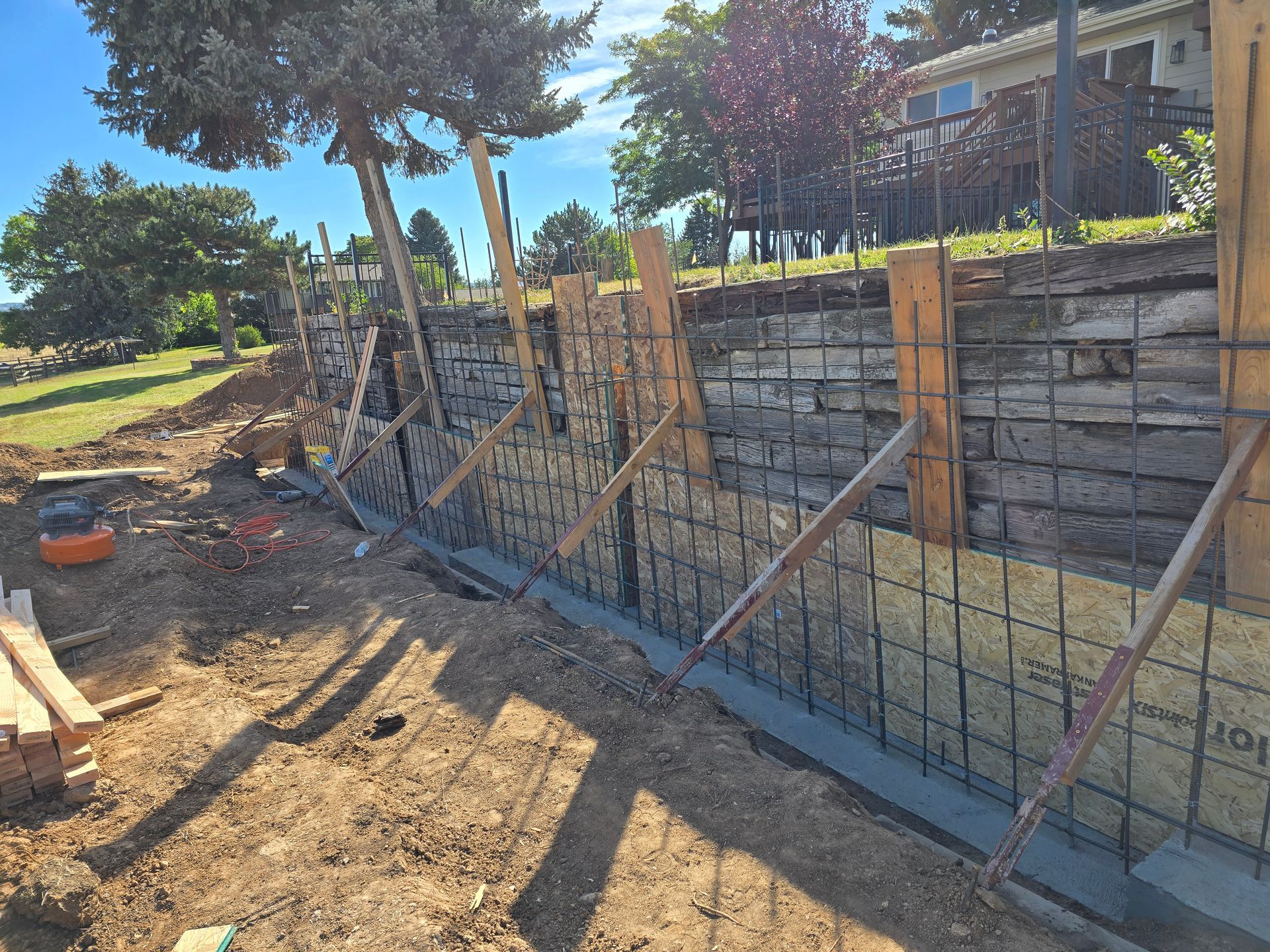 Construction site with rebar foundation forms along a stone wall beside trees and a building