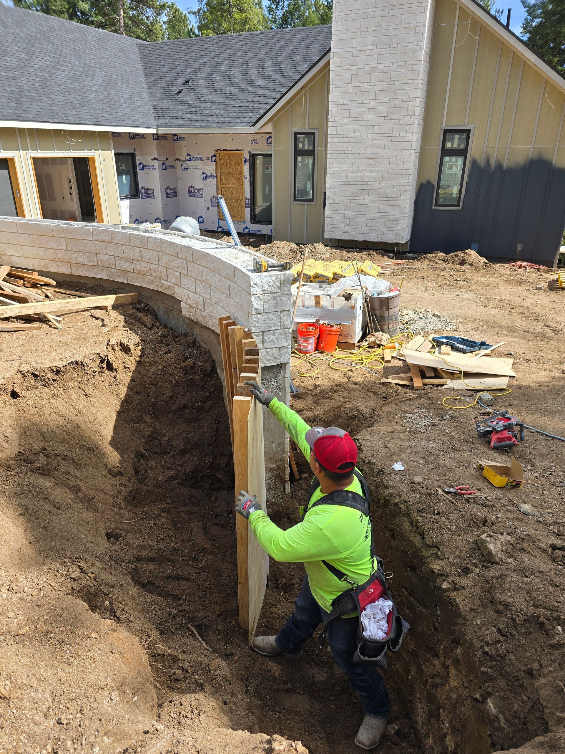 Worker in neon green shirt and red cap bracing a retaining wall trench at a house construction site