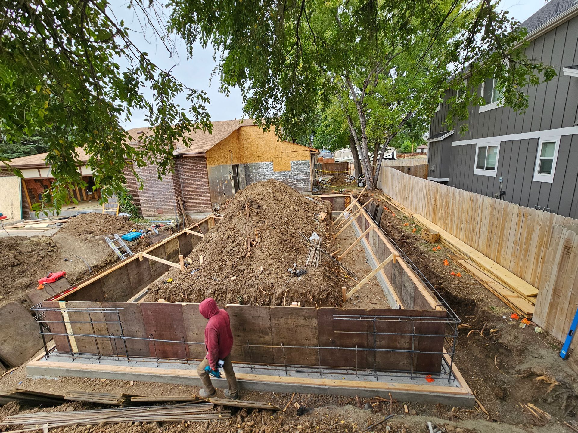 Construction site with a large dug trench, dirt piles, and a worker standing by a fenced excavation.