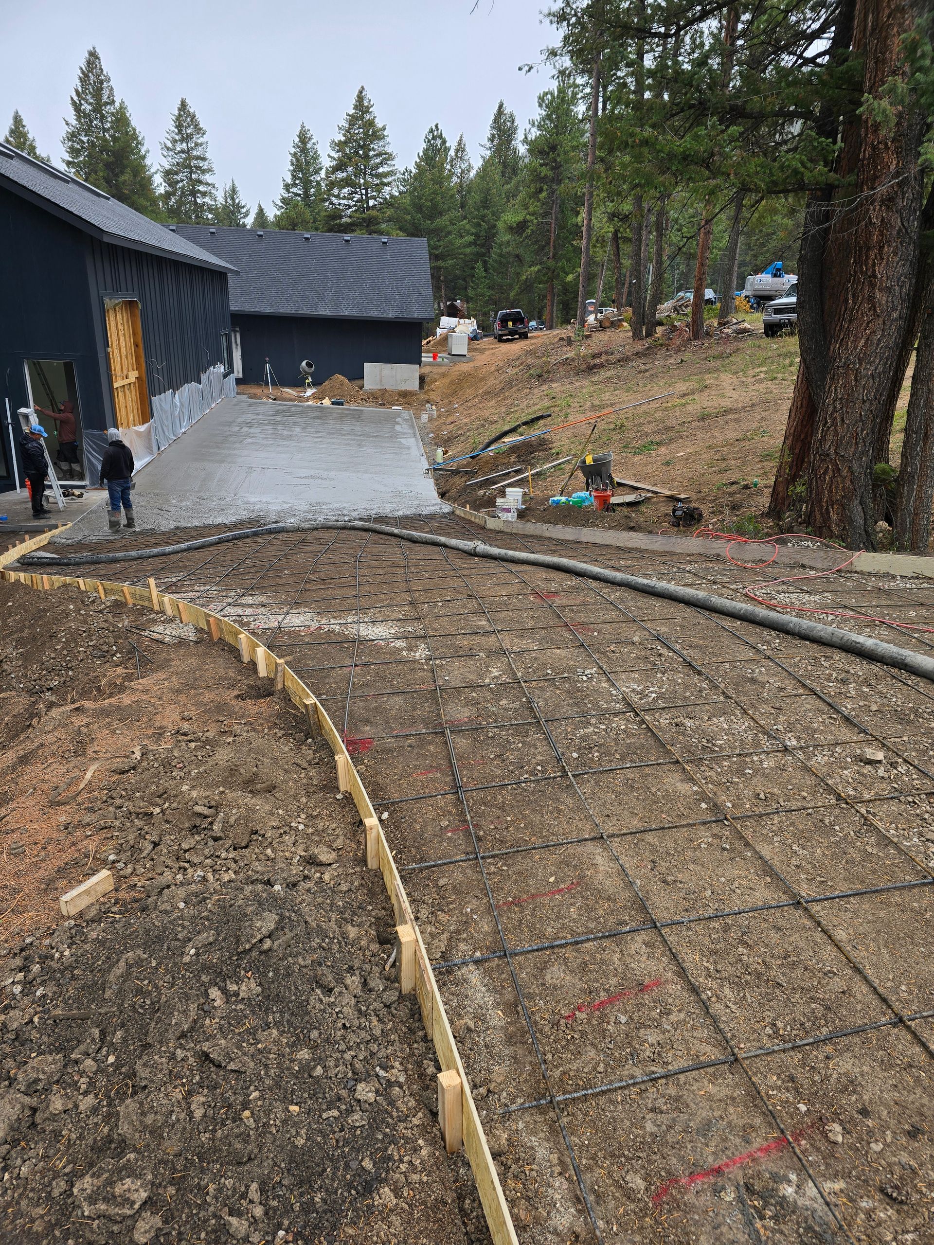 Gravel path curving beside a dark house on a wooded hillside, with trees and construction materials nearby