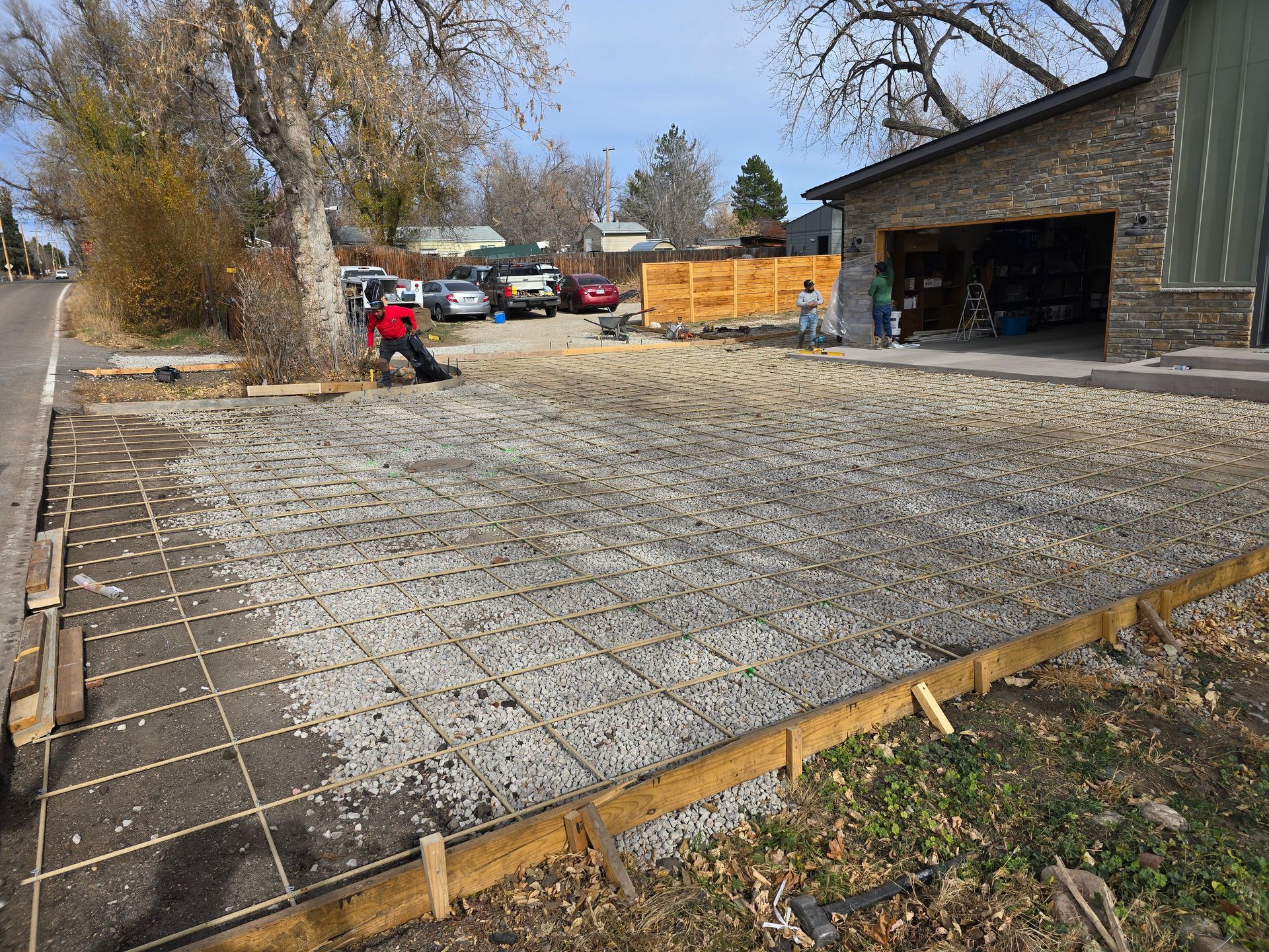 Worker smoothing concrete driveway beside a stone house and garage, with wooden forms and gravel in a suburban yard