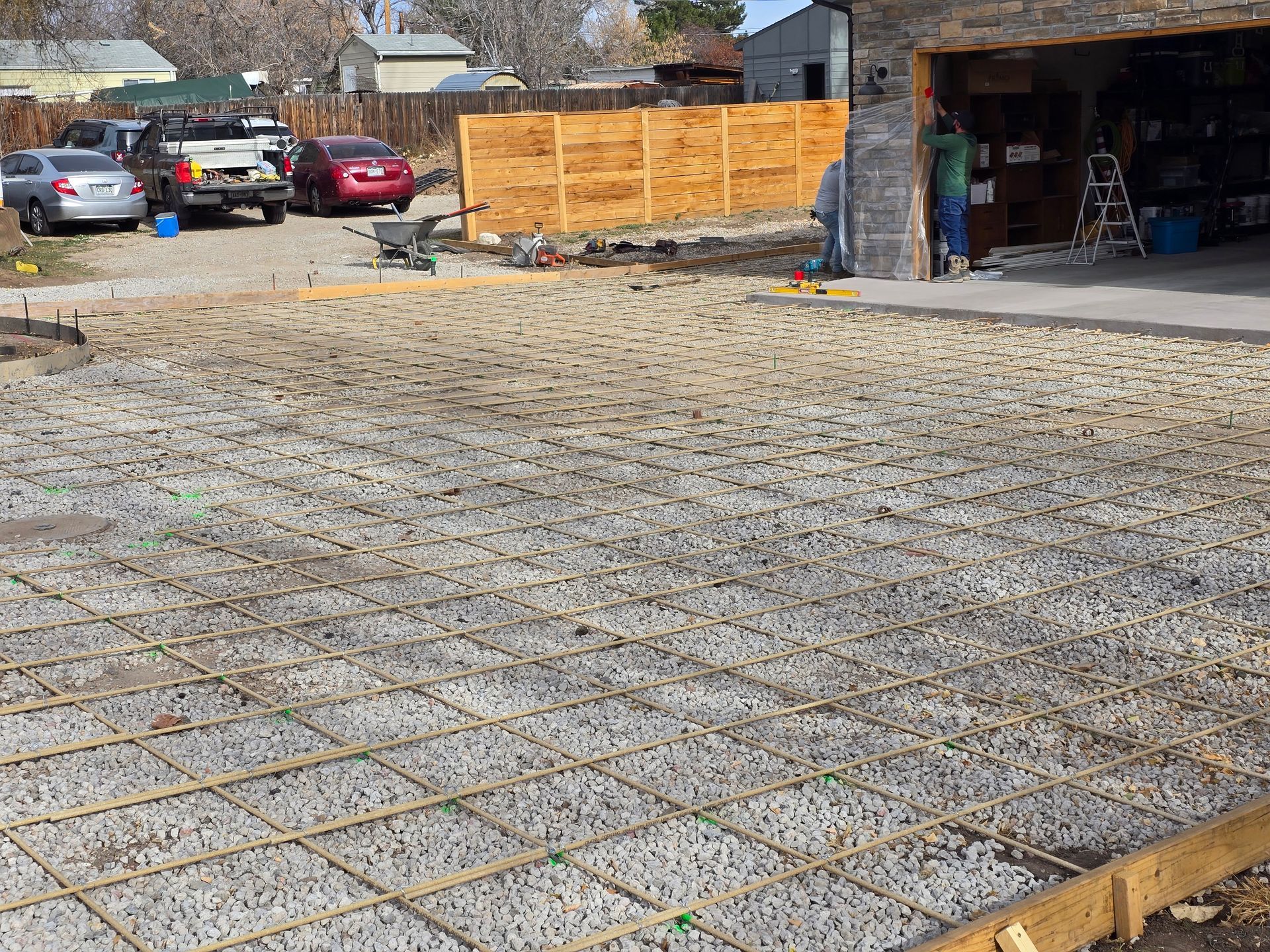Gravel foundation with rebar grid at a construction site beside a wooden fence and open garage