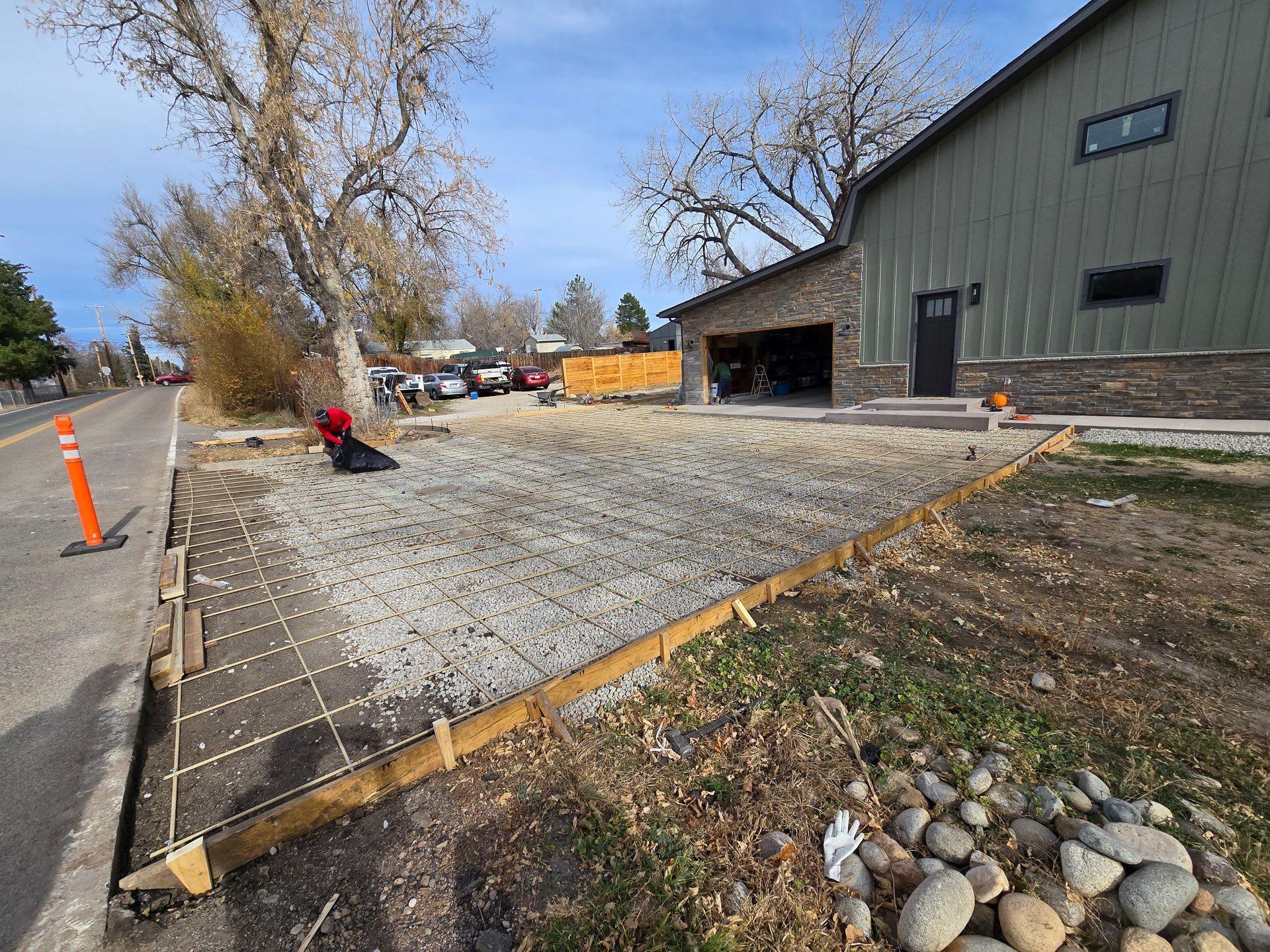 Gravel construction area beside a road and green building, with orange cones and a worker near excavation.