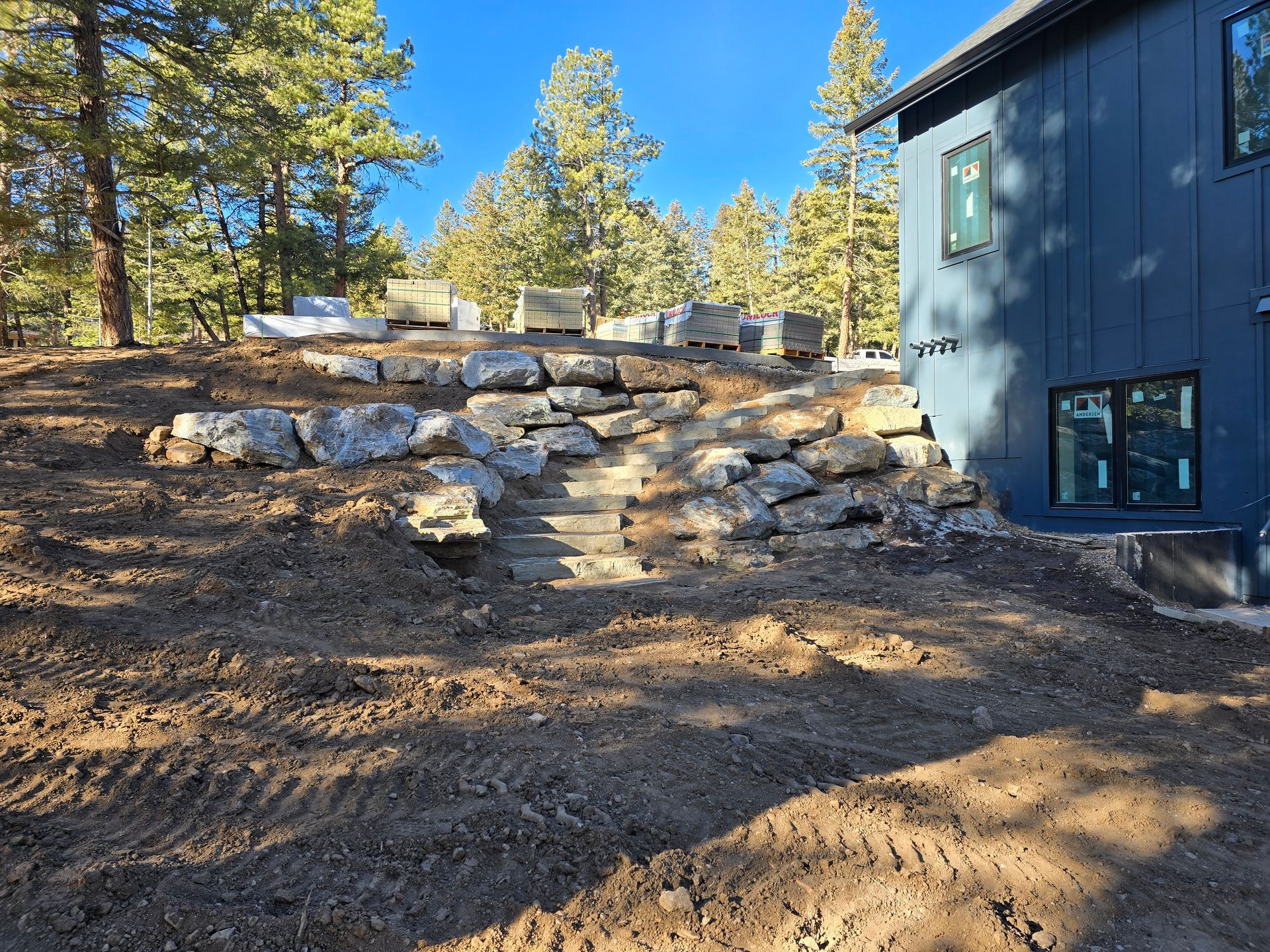 Stone steps in a wooded yard beside a blue house, with pine trees and stacked stones in the background.