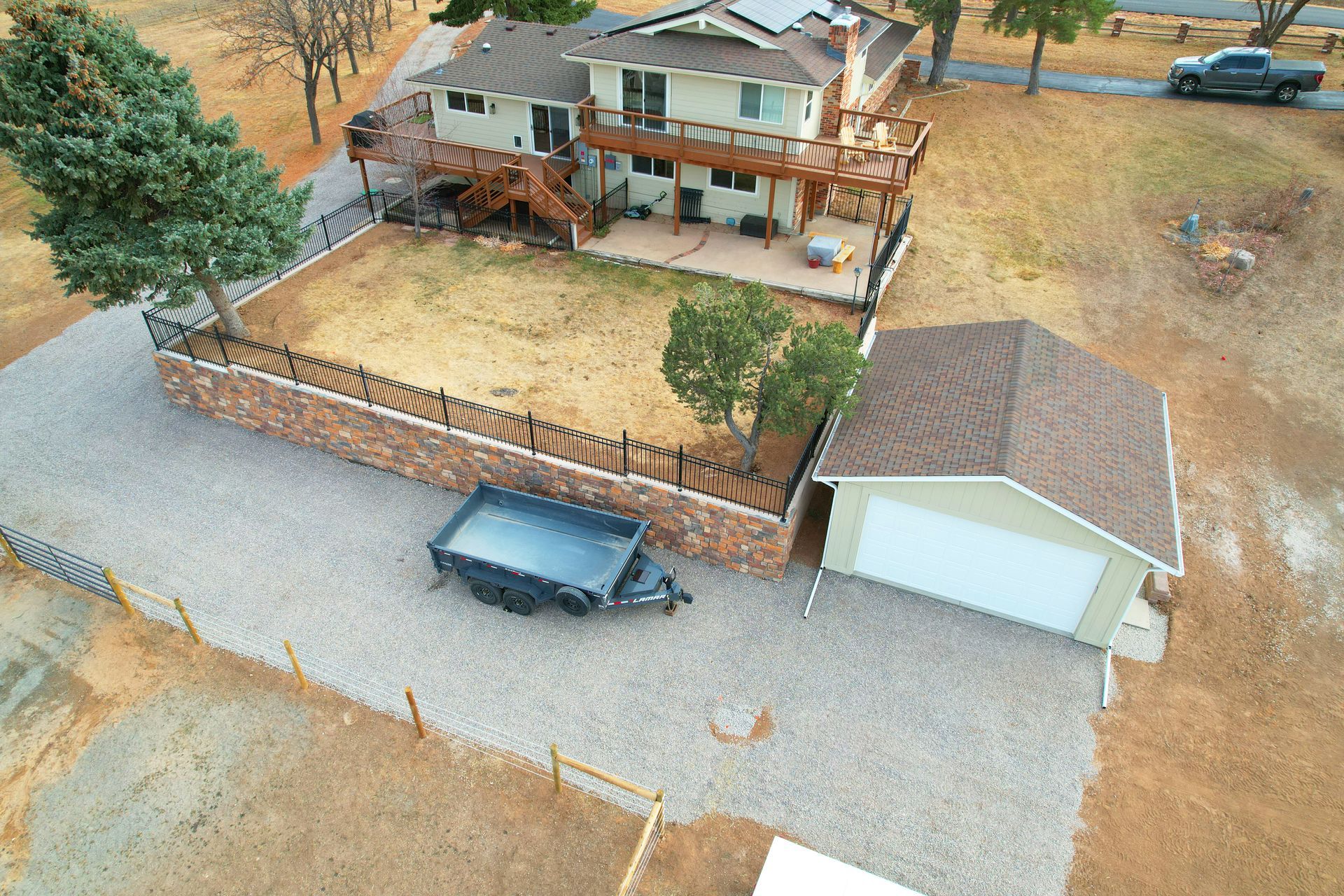 Aerial view of a house with a fenced yard, detached garage, and parked vehicles on a rural lot.