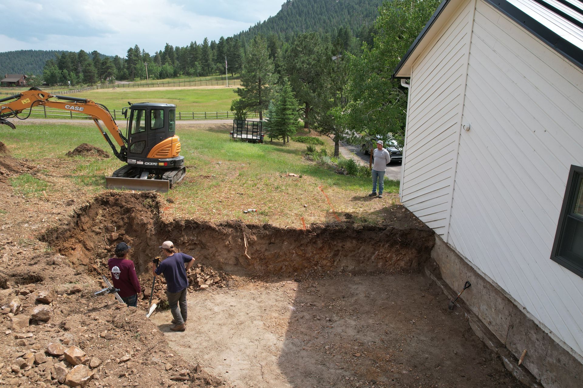 Workers near an excavated foundation beside a white house, with an excavator and rural mountains in the background