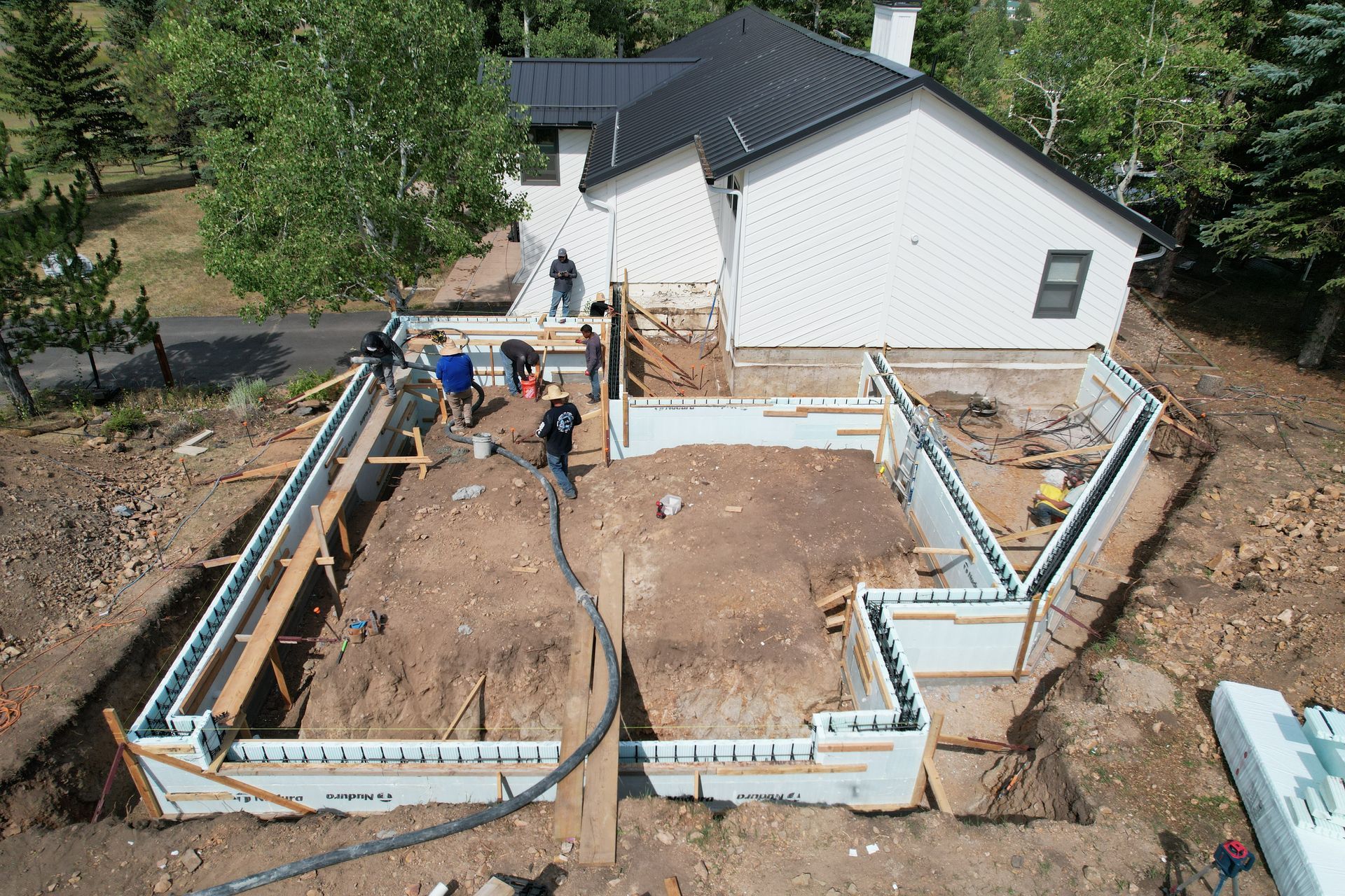 Construction crew setting concrete foundation forms beside a white house and deck.