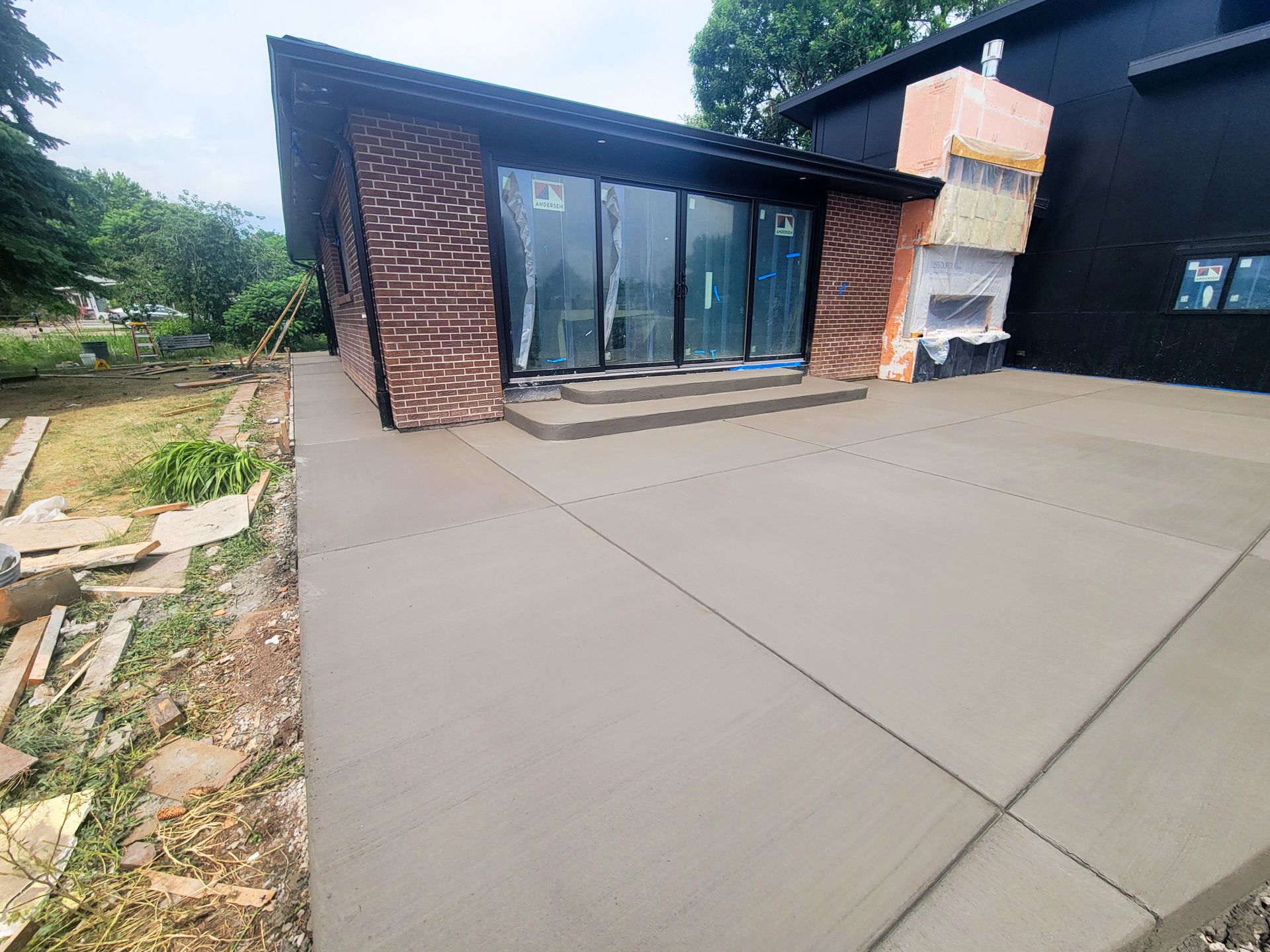 Modern house exterior with large concrete patio and glass doors, surrounded by greenery