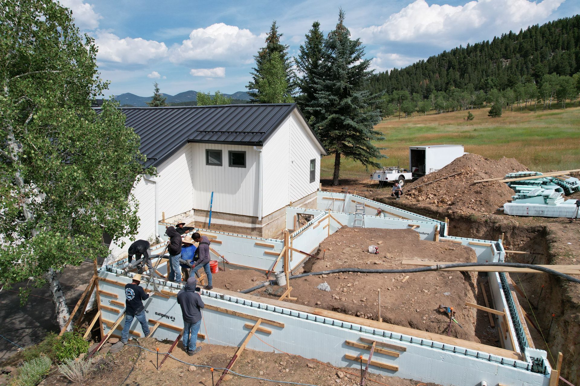 Workers building a house foundation beside a white home on a dirt lot with trees and mountains nearby