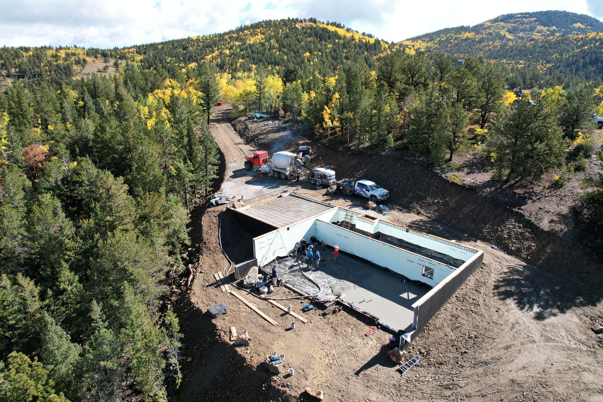 Aerial view of a remote construction site with a concrete pool, canopy, vehicles, and forested hills.