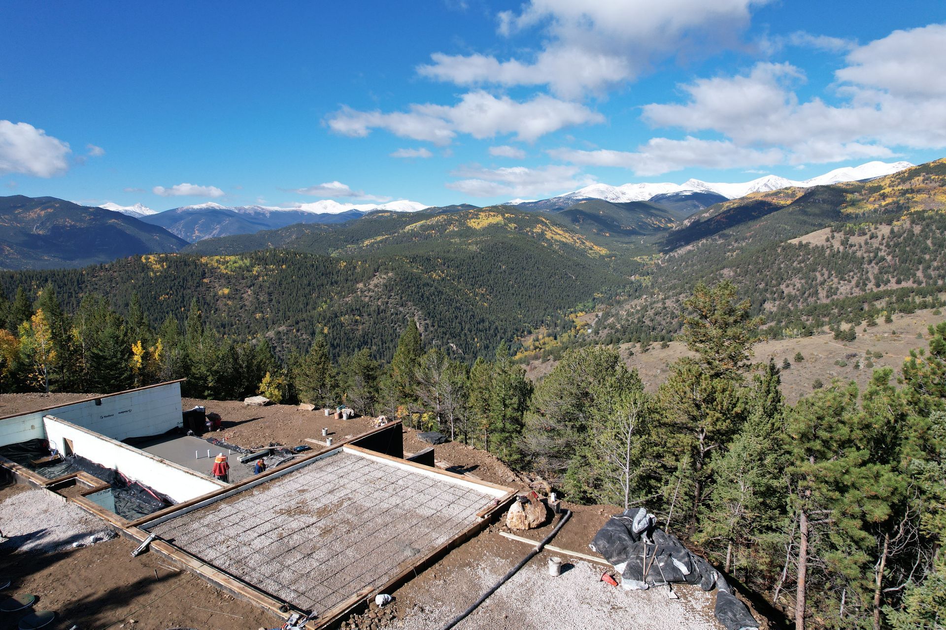 Mountain valley view from a rooftop, with pine-covered hills, distant peaks, and blue sky with clouds.