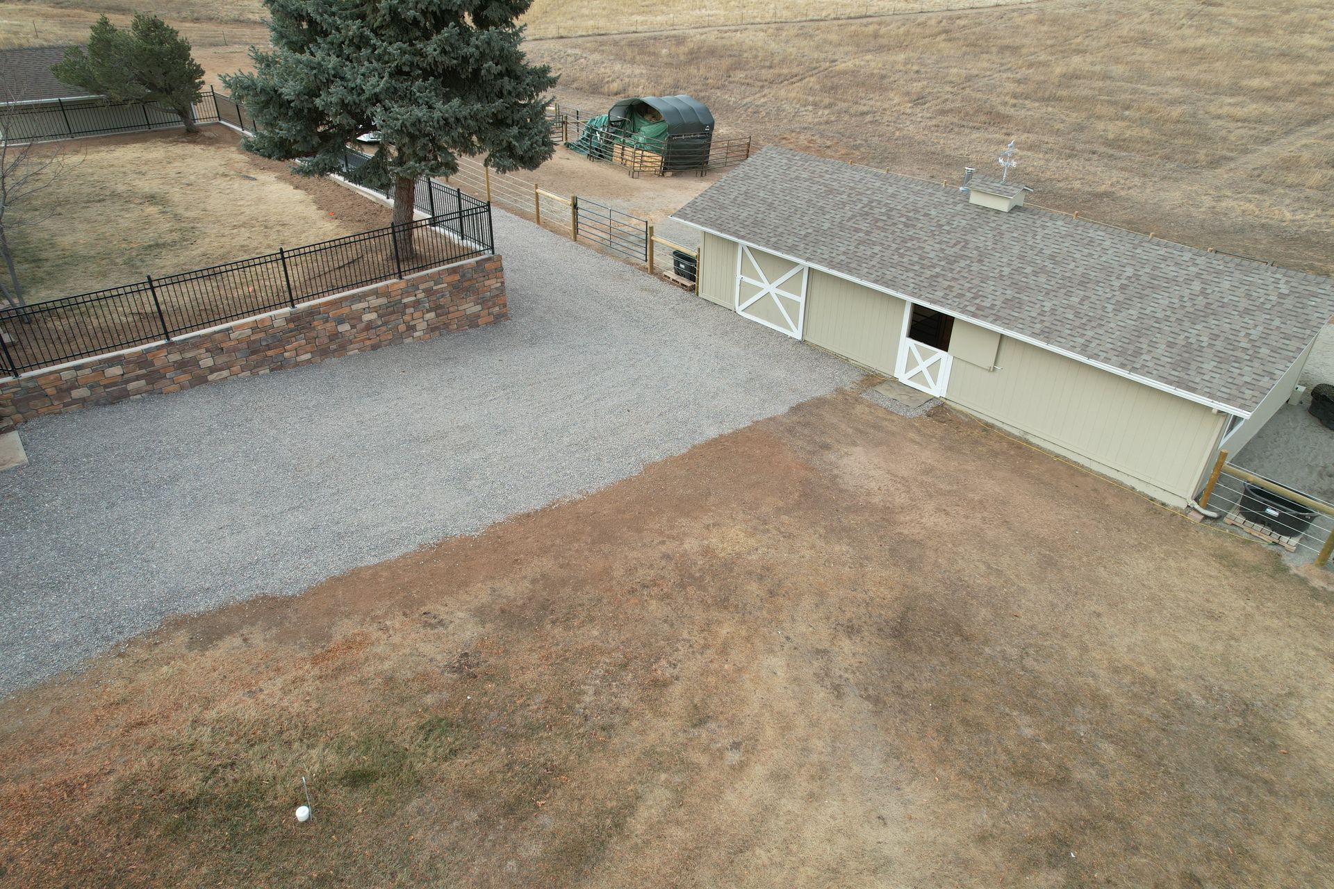 Gravel driveway beside a beige house with a brown roof and a fenced yard in a rural setting