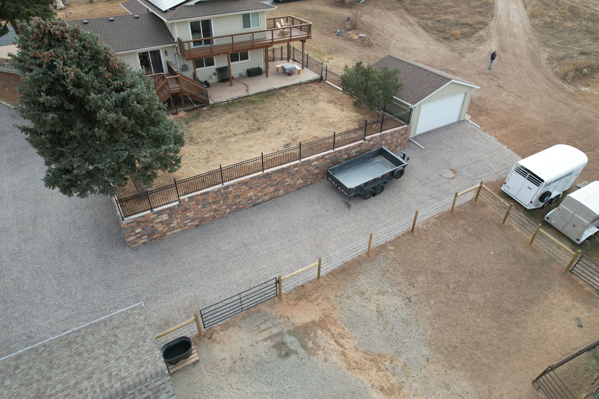 Aerial view of a rural house with a gravel driveway, stone wall, parked truck, van, and a large tree.