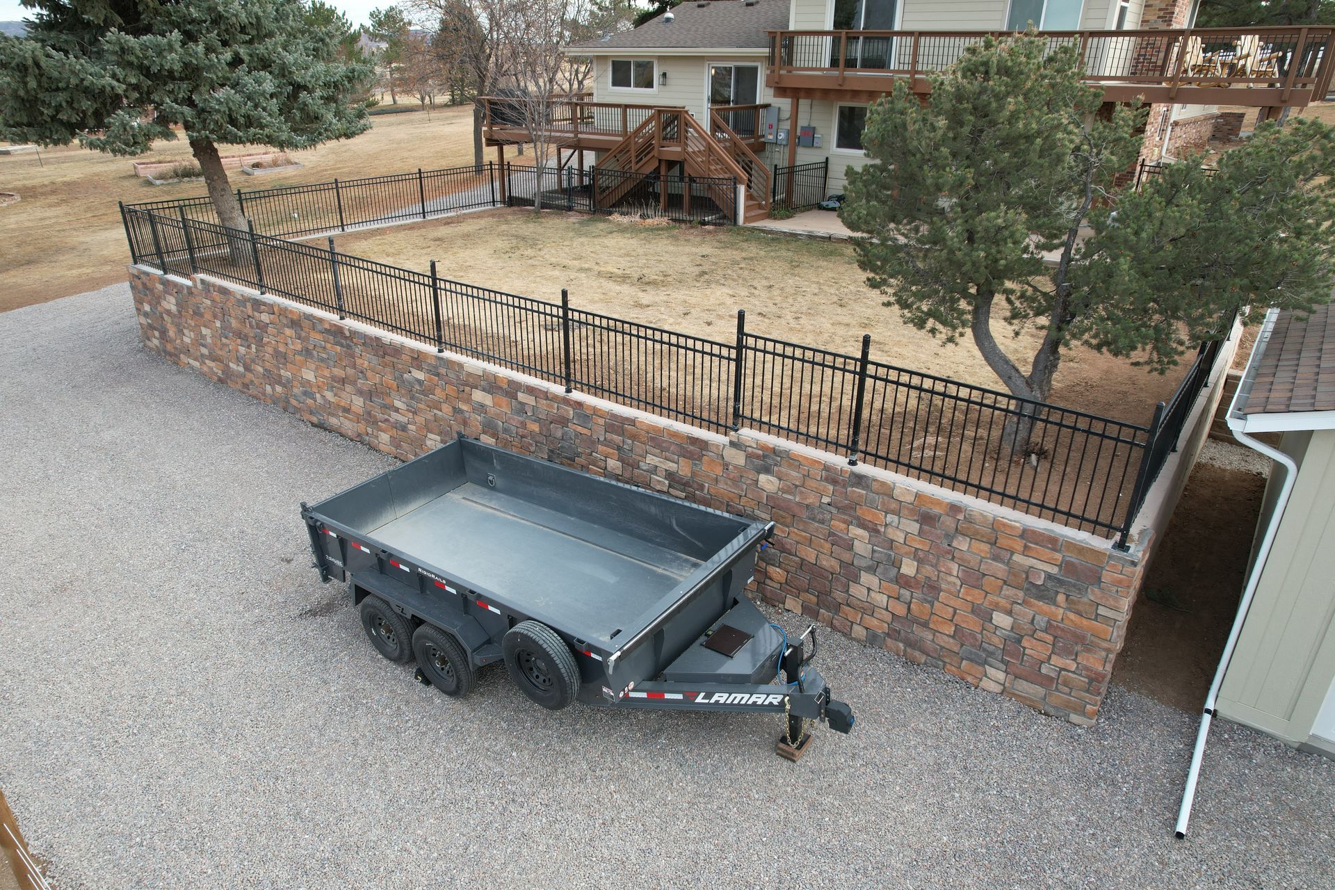 Black utility trailer on gravel beside a stone fence and house patio with outdoor furniture.