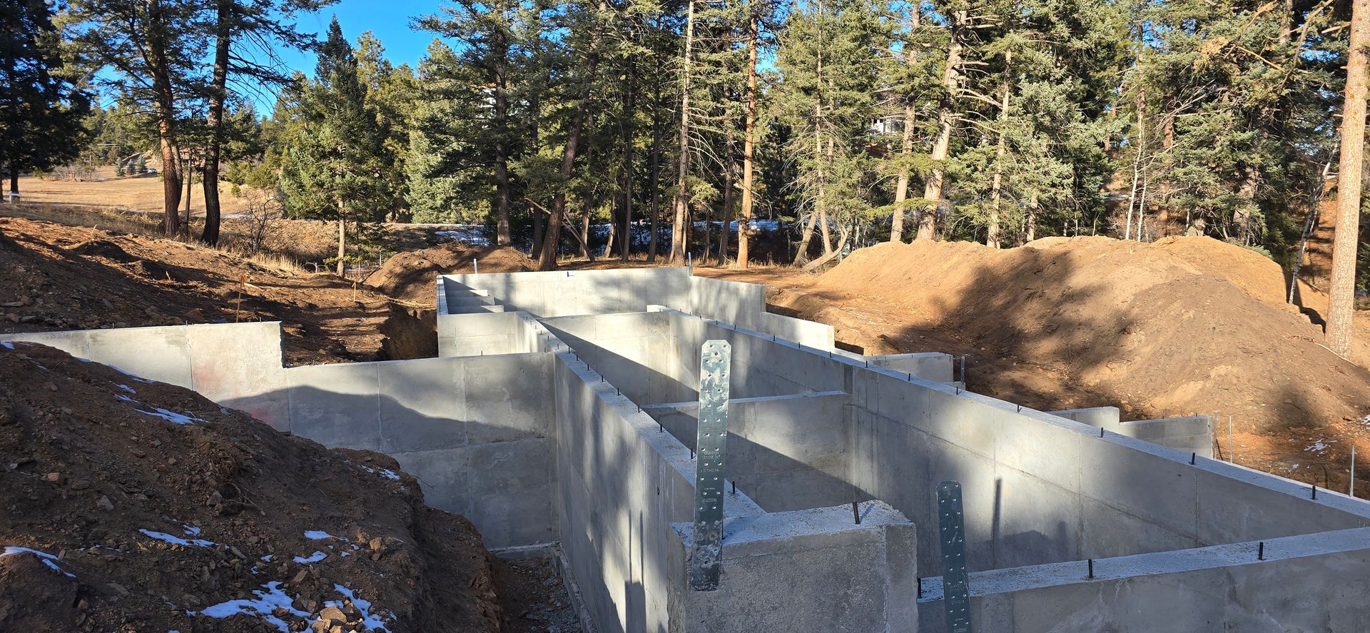 Concrete foundation walls under construction in a wooded area with dirt and pine trees.