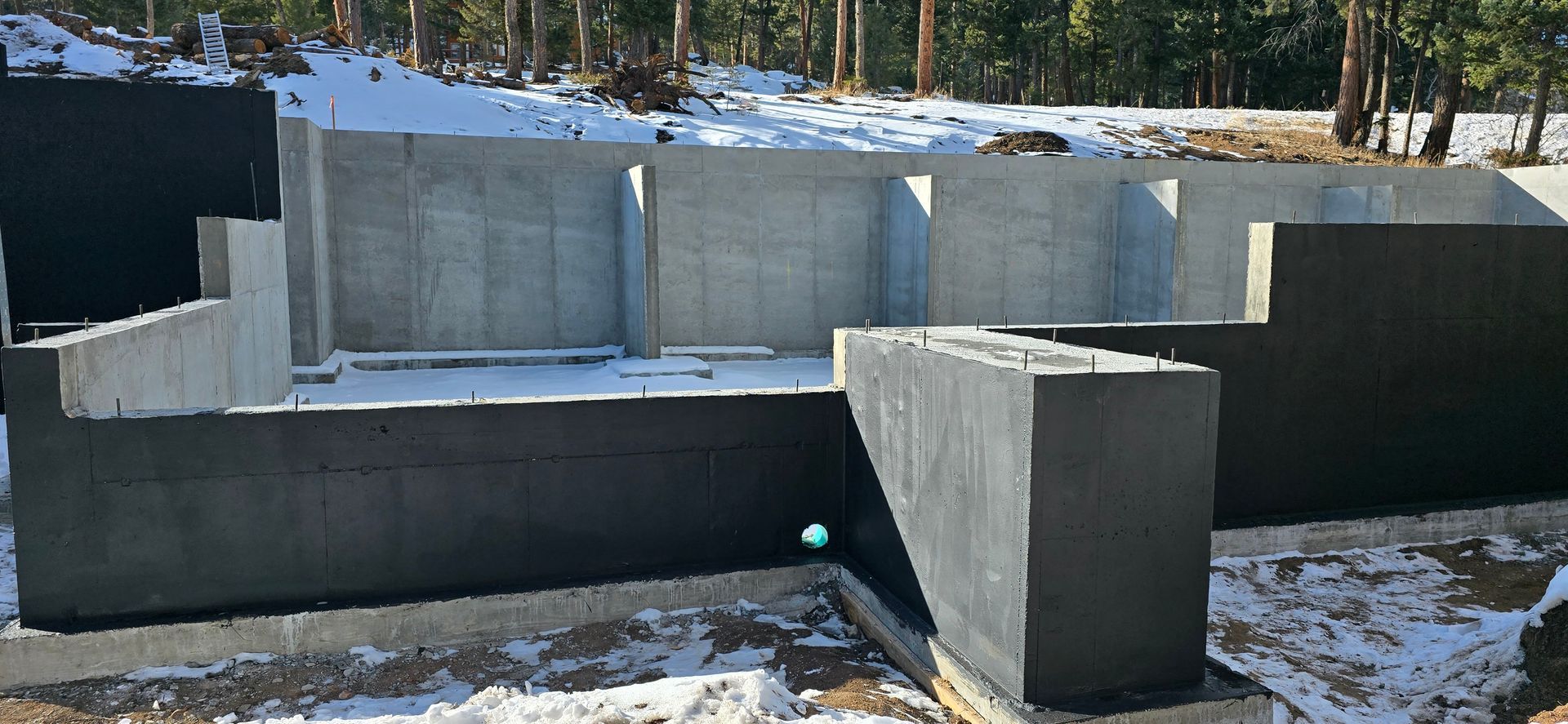 Concrete foundation walls under construction in a snowy outdoor site with wooded hills in the background
