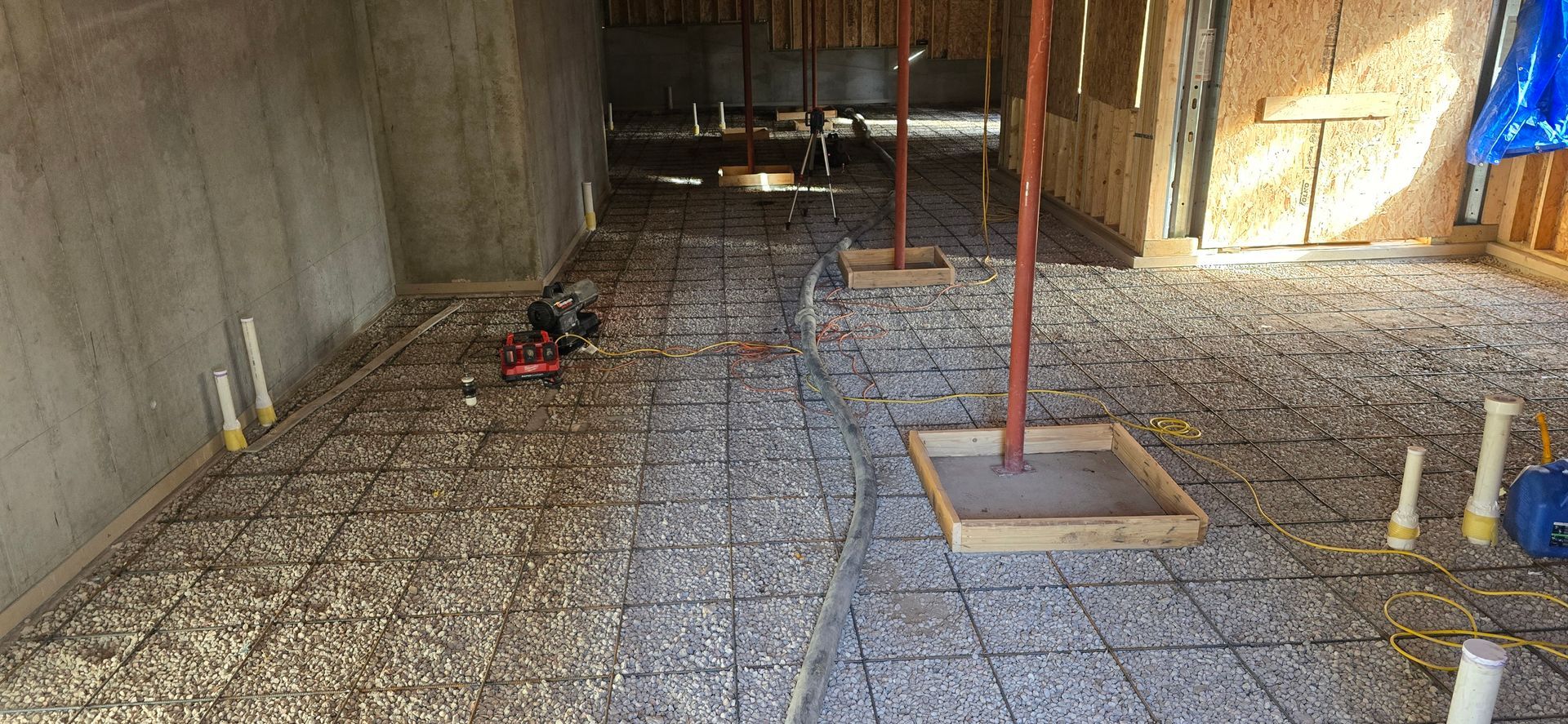 Interior construction site with rebar grid, concrete forms, and red support posts on a dirt floor