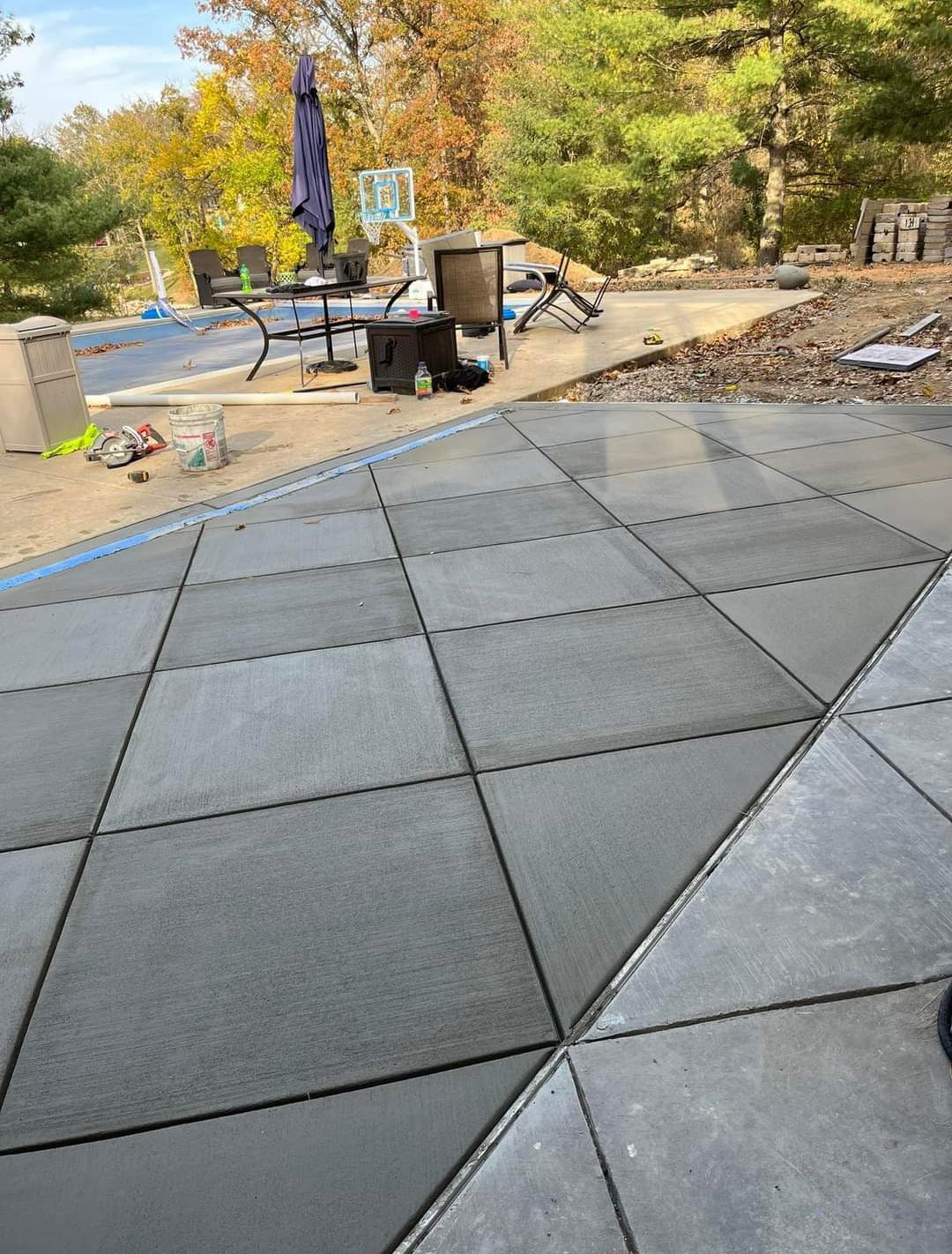 Outdoor patio with gray pavers, a table, chairs, and a closed umbrella in a leafy backyard