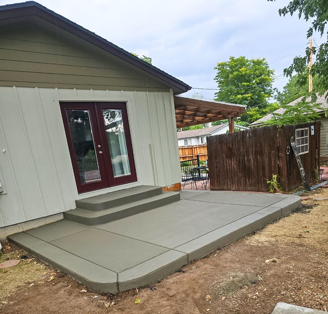 Concrete patio and steps beside a white house with maroon-trimmed windows and a wooden fence