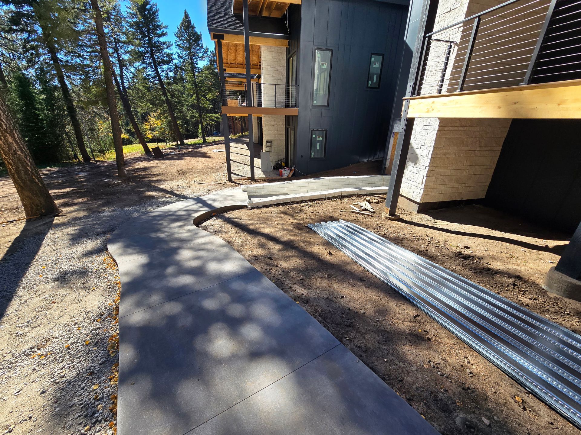 Curved concrete walkway beside a modern house and wooden deck in a wooded area, with construction materials nearby